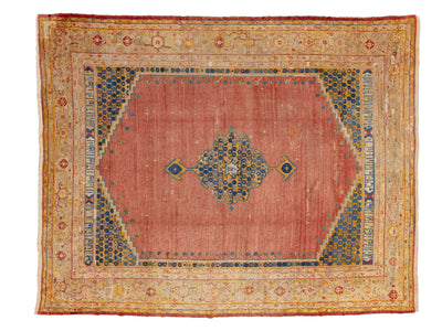 A rectangular antique area rug with a dusty rose pink center field, a blue and gold central medallion, and a wide beige and gold ornamental border.