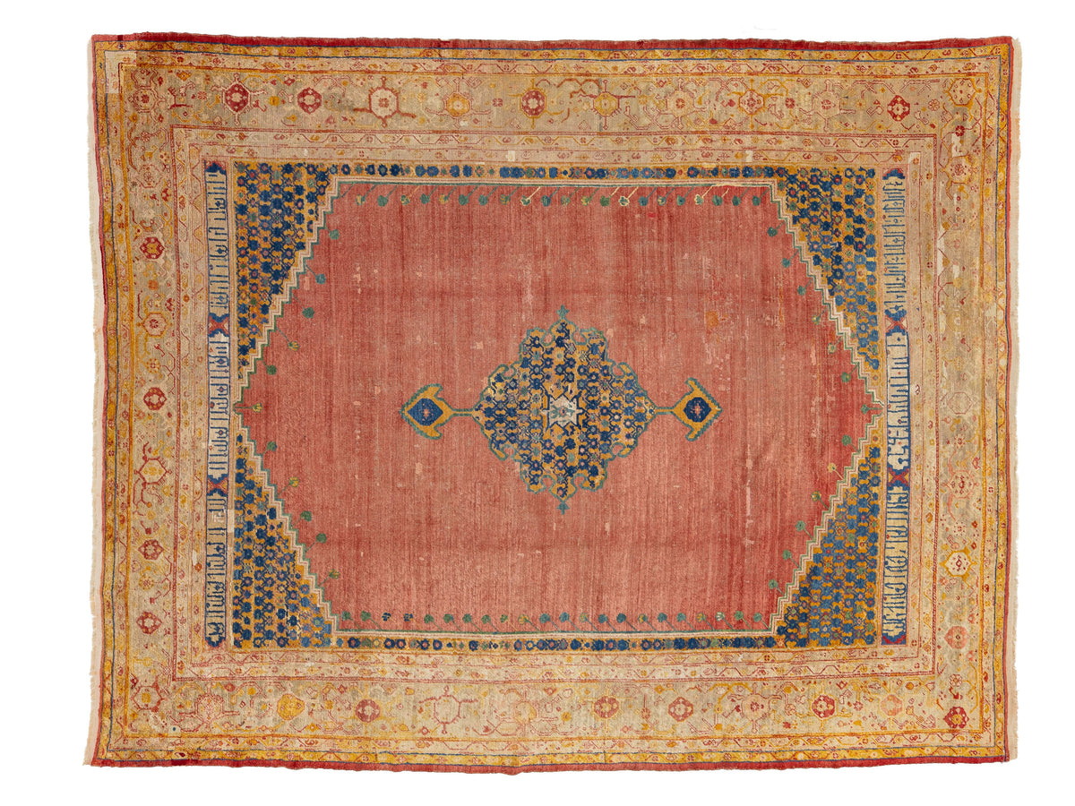 A rectangular antique area rug with a dusty rose pink center field, a blue and gold central medallion, and a wide beige and gold ornamental border.