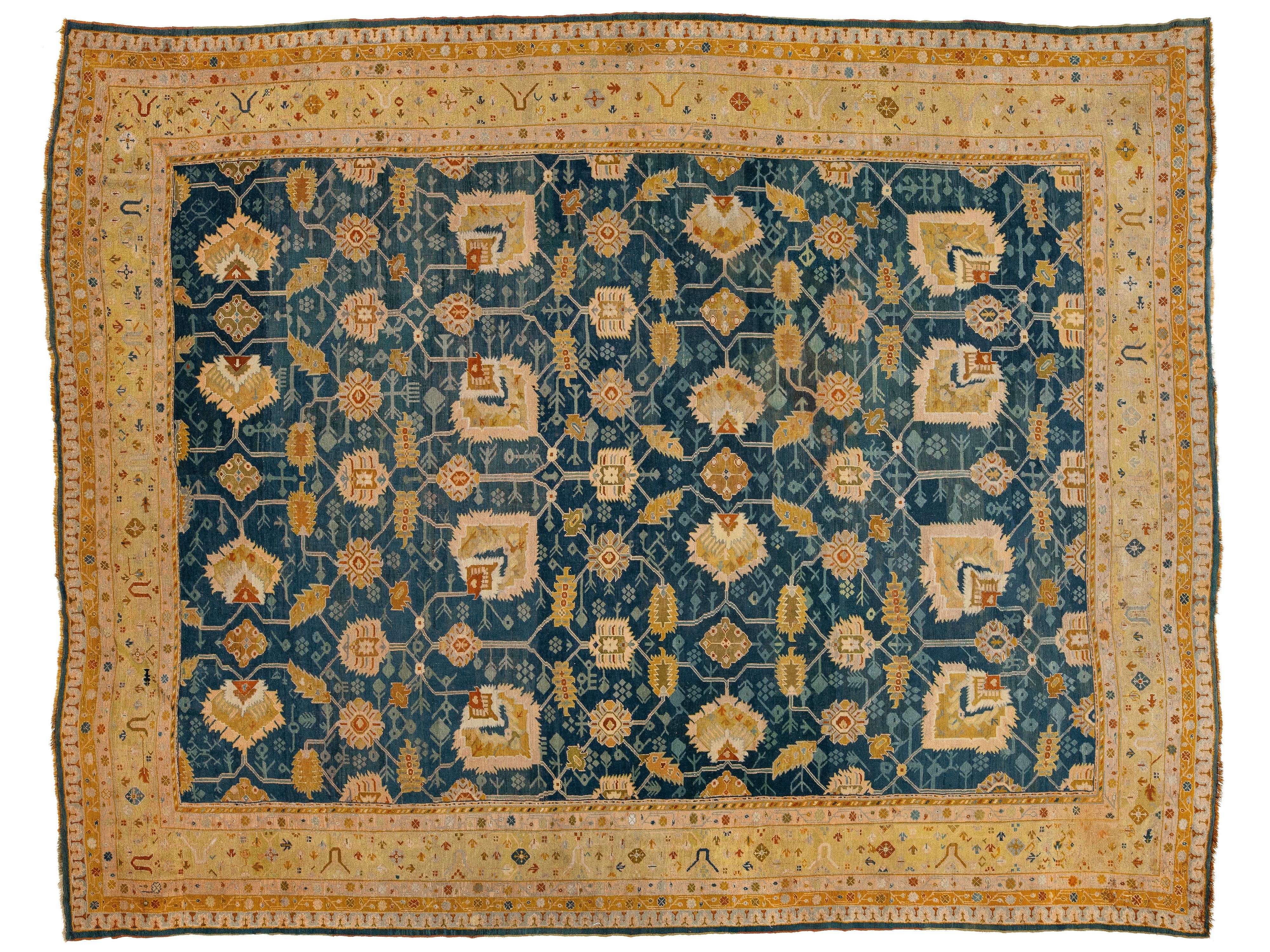A rectangular traditional oriental rug with a deep teal blue field, decorated with a repeating cream and gold floral pattern, surrounded by a wide cream border with intricate gold and red details.