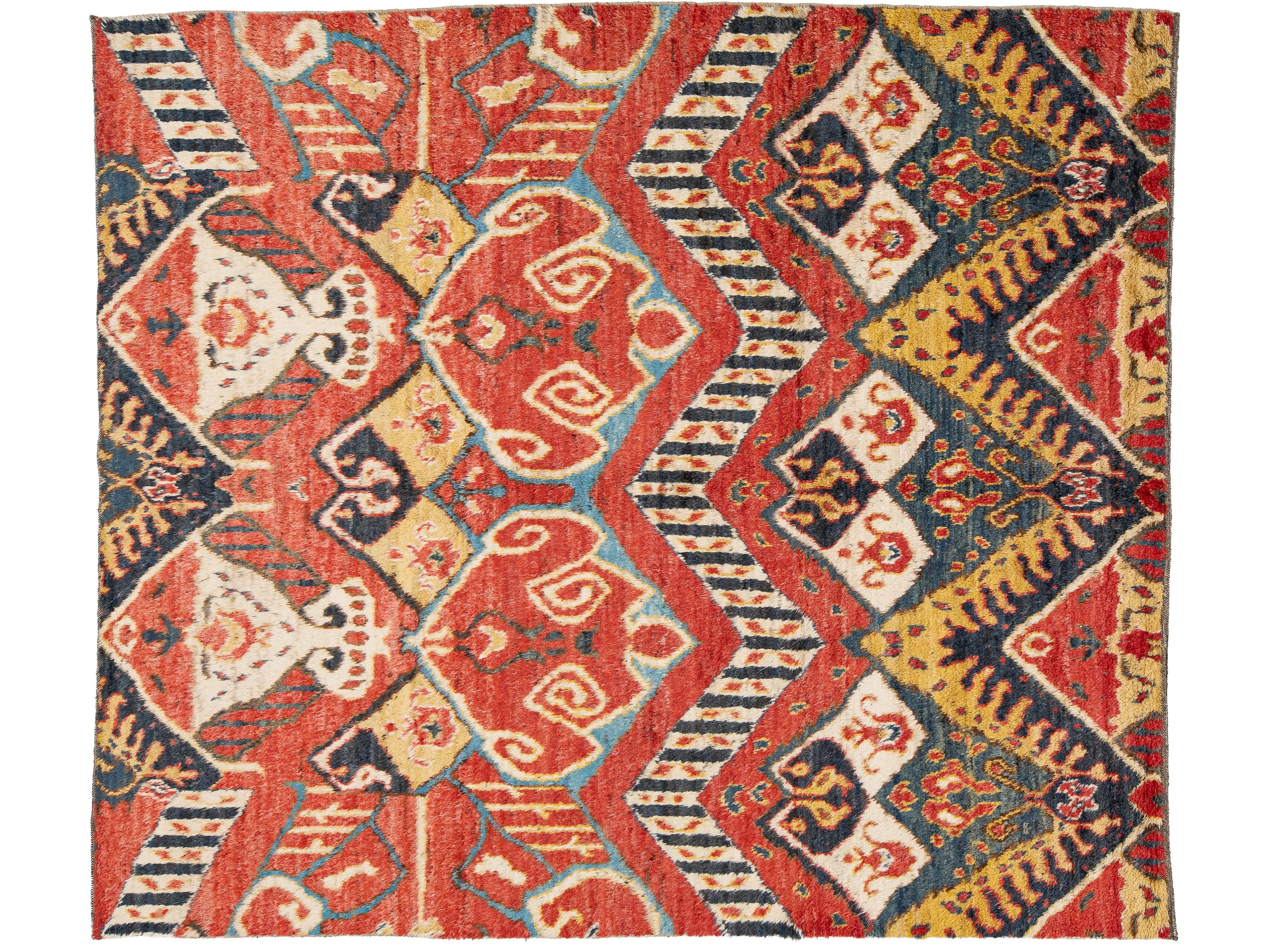 A rectangular area rug with a complex geometric and tribal pattern featuring red, navy blue, cream, and mustard yellow.