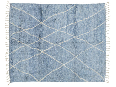 A rectangular blue shag rug with a white geometric crisscross pattern and white fringe tassels on all four sides, shown flat against a white background.