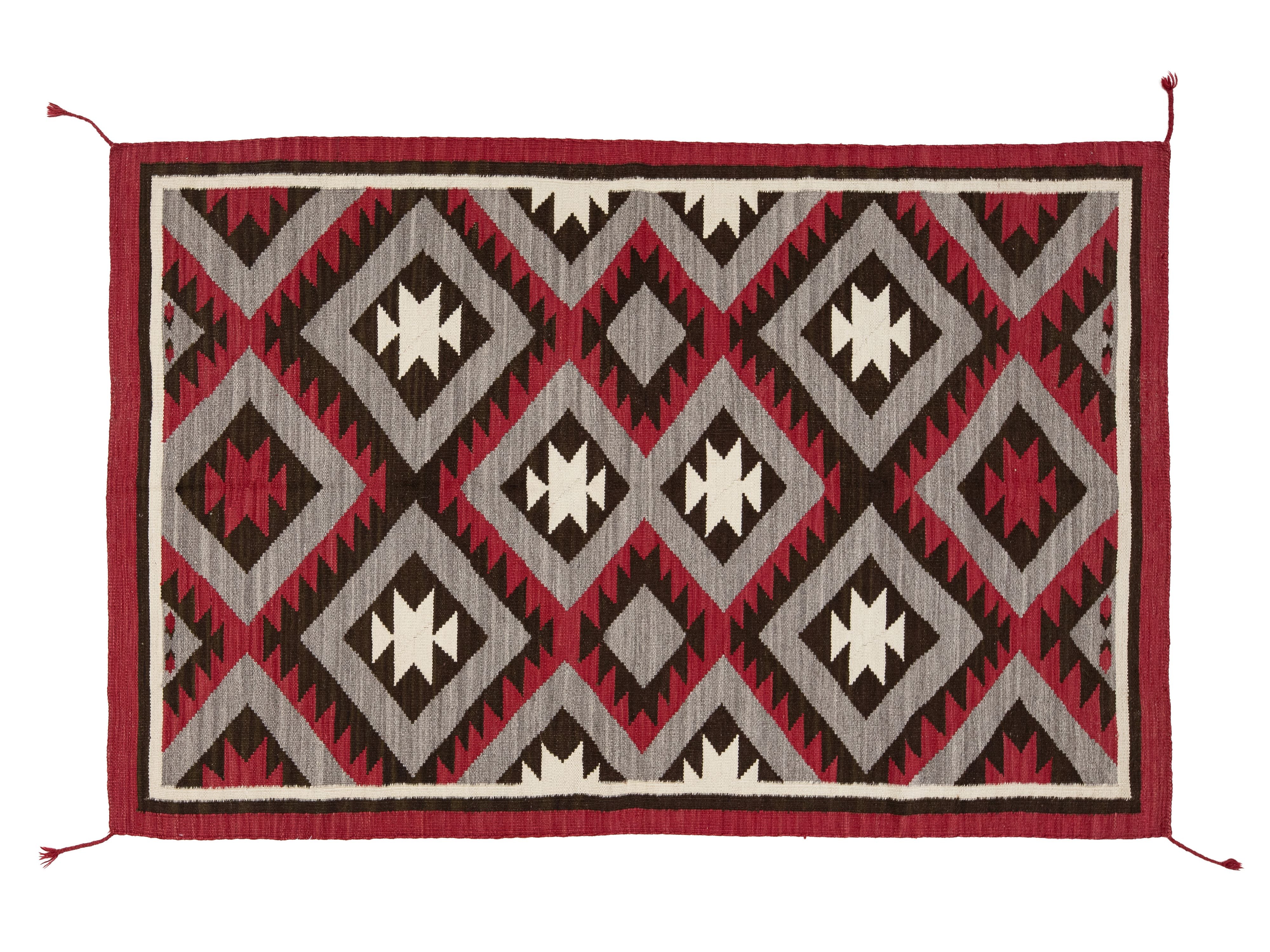A rectangular woven rug with a Southwestern geometric pattern in red, dark brown, light gray, and off-white, featuring a solid red border and corner tassels on a white background.