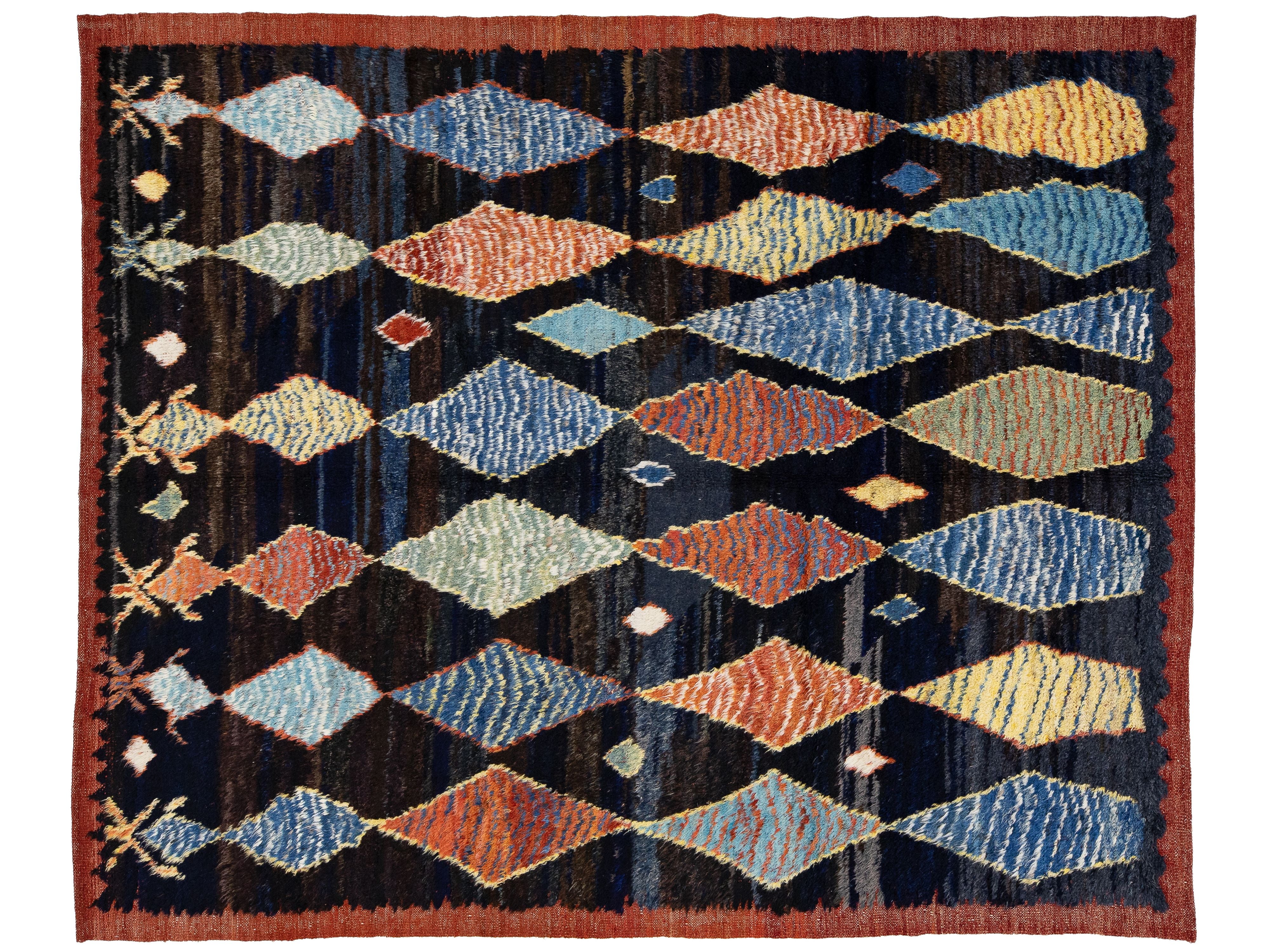 A rectangular woven rug with a dark navy and black background, featuring a repeating pattern of stylized diamond shapes in muted blue, rust orange, beige, and yellow, finished with a rust-colored border.