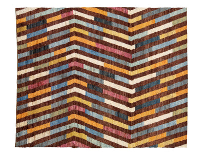 A rectangular woven textile or rug with a bold chevron pattern made of horizontal multicolored stripes in brown, cream, yellow, blue, pink, and gray.