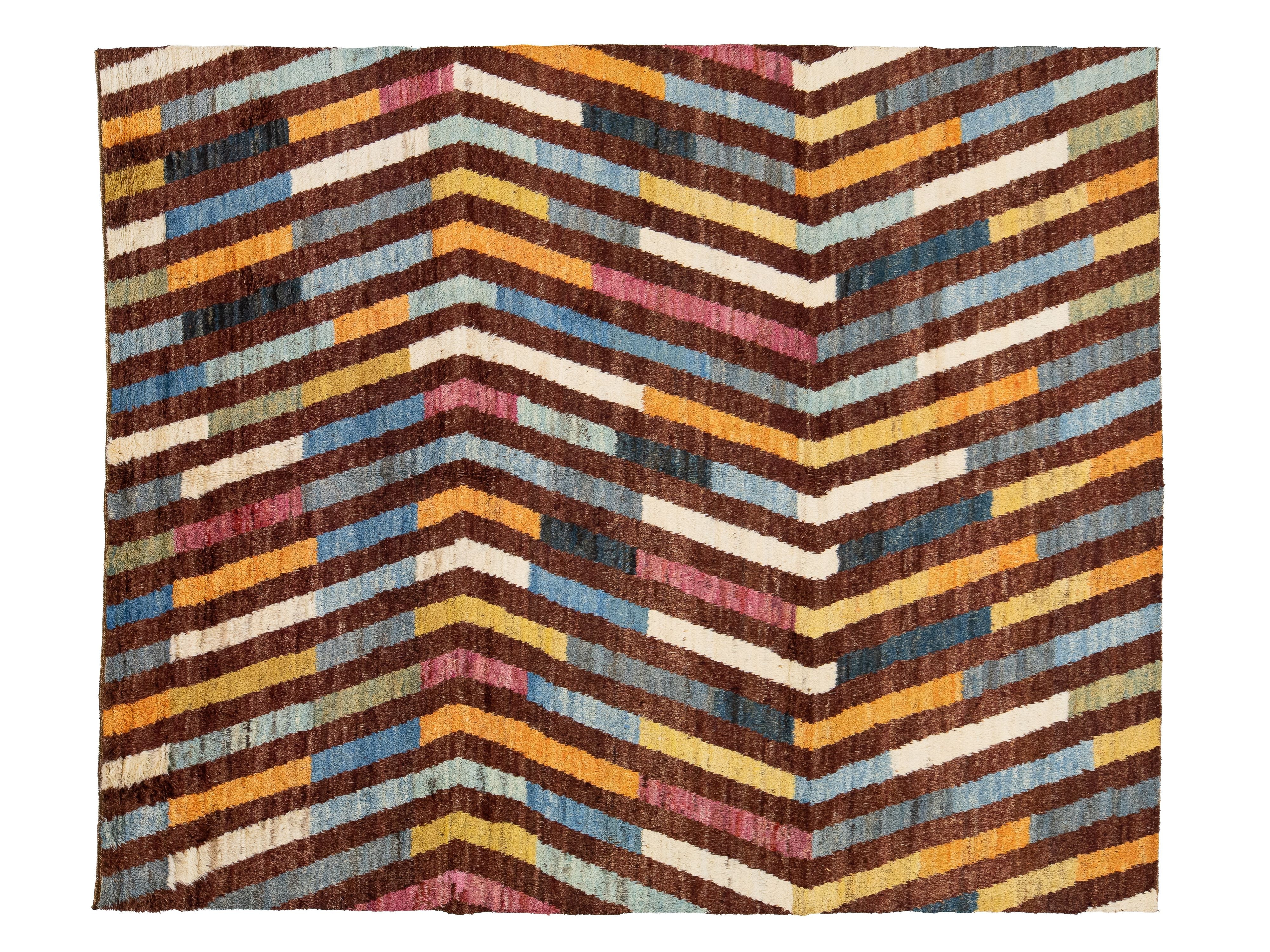 A rectangular woven textile or rug with a bold chevron pattern made of horizontal multicolored stripes in brown, cream, yellow, blue, pink, and gray.