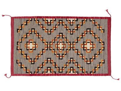 A rectangular woven rug with a geometric Southwestern pattern, featuring a gray background with red, orange, black, and cream motifs, finished with a red border and corner tassels.