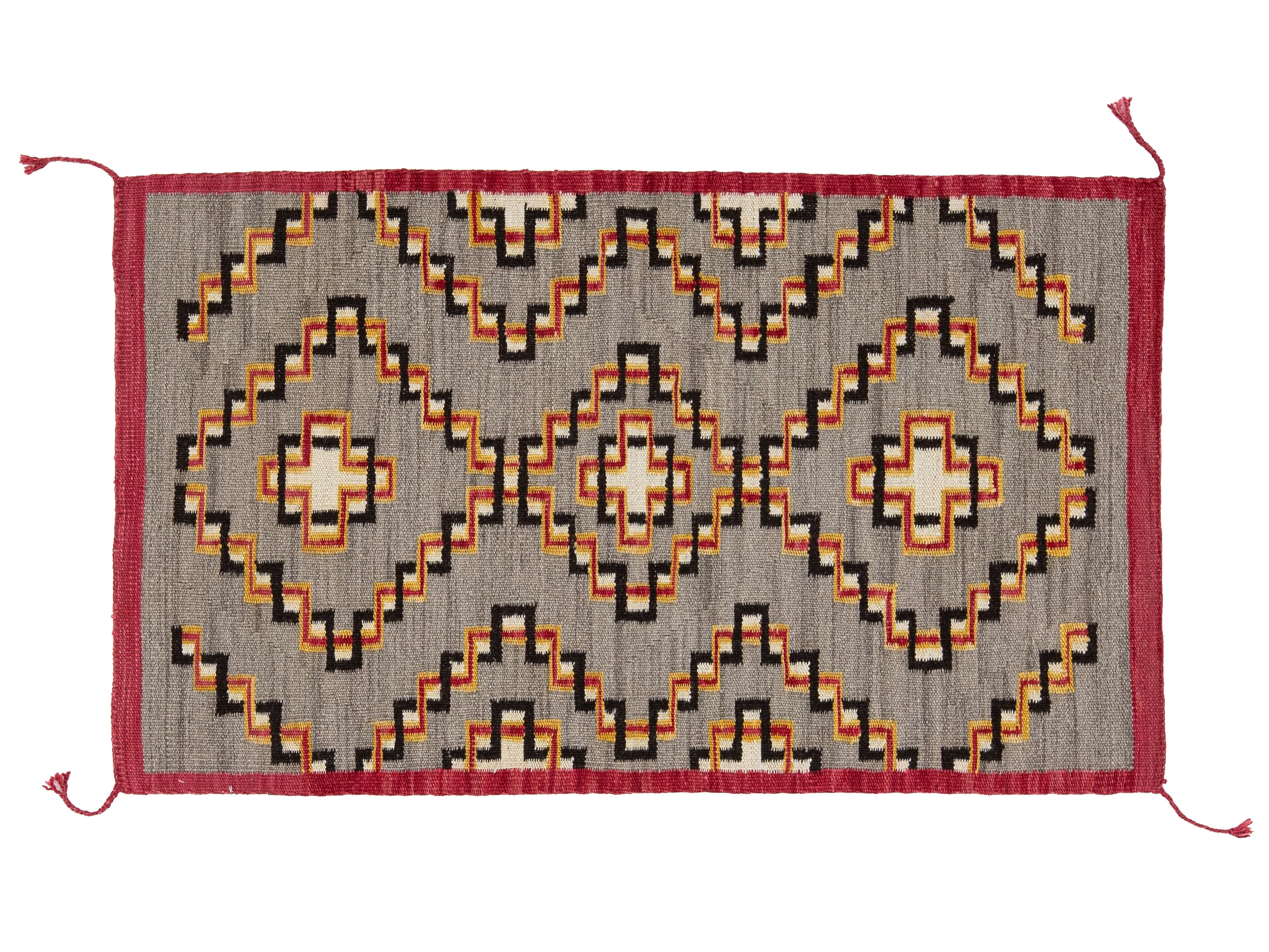 A rectangular woven rug with a geometric Southwestern pattern, featuring a gray background with red, orange, black, and cream motifs, finished with a red border and corner tassels.