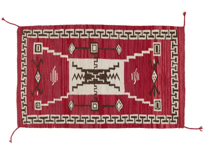 A rectangular woven rug with a red field, cream central panel, dark brown geometric motifs, and a repeating border pattern, shown flat against a white background.