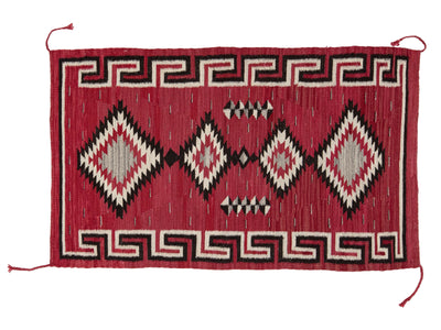 A rectangular woven rug with a red field, featuring large diamond motifs in black, cream, and red, bordered by geometric patterns.