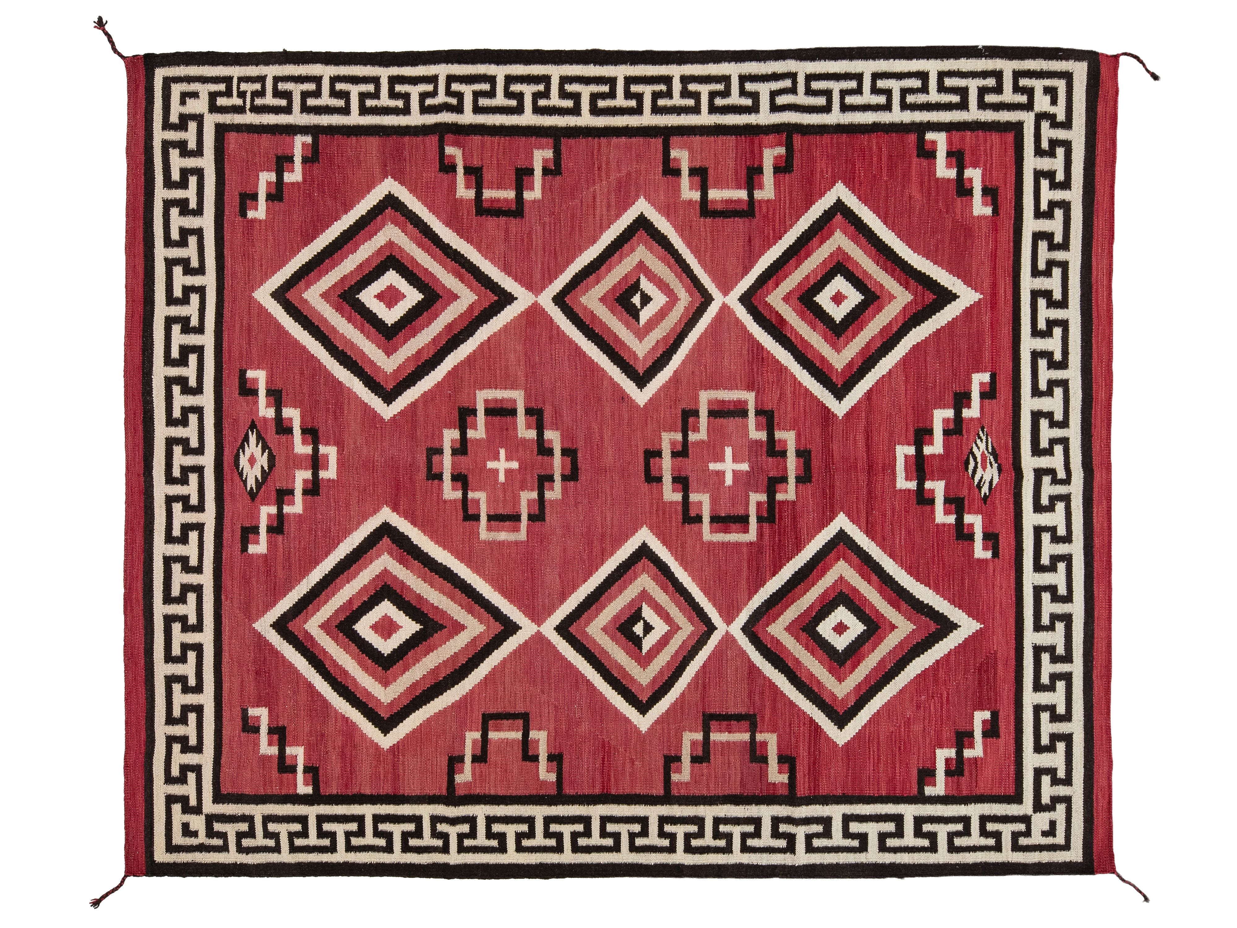 A rectangular woven rug with a red field, black and cream geometric diamond motifs, and a black Greek key border.