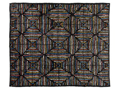 A rectangular woven rug with a bold geometric pattern of nested squares and stripes in black, blue, red, yellow, and white, presented flat against a white background.