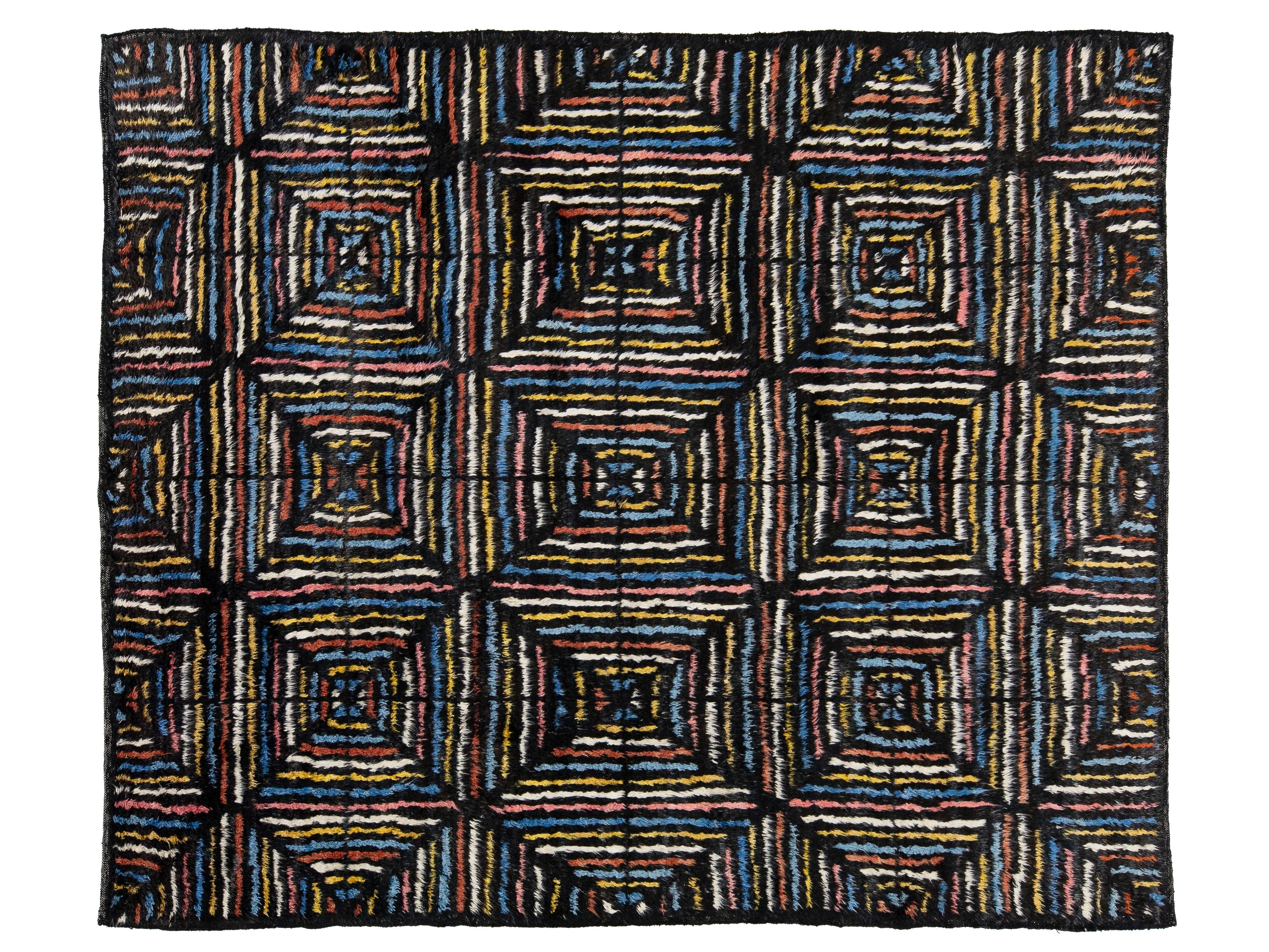 A rectangular woven rug with a bold geometric pattern of nested squares and stripes in black, blue, red, yellow, and white, presented flat against a white background.