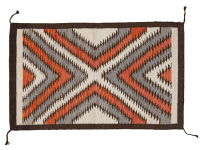 A rectangular woven textile rug or mat with a geometric Southwestern-style pattern in rust orange, gray, black, and off-white, finished with a dark brown border and hanging tassels at the corners.