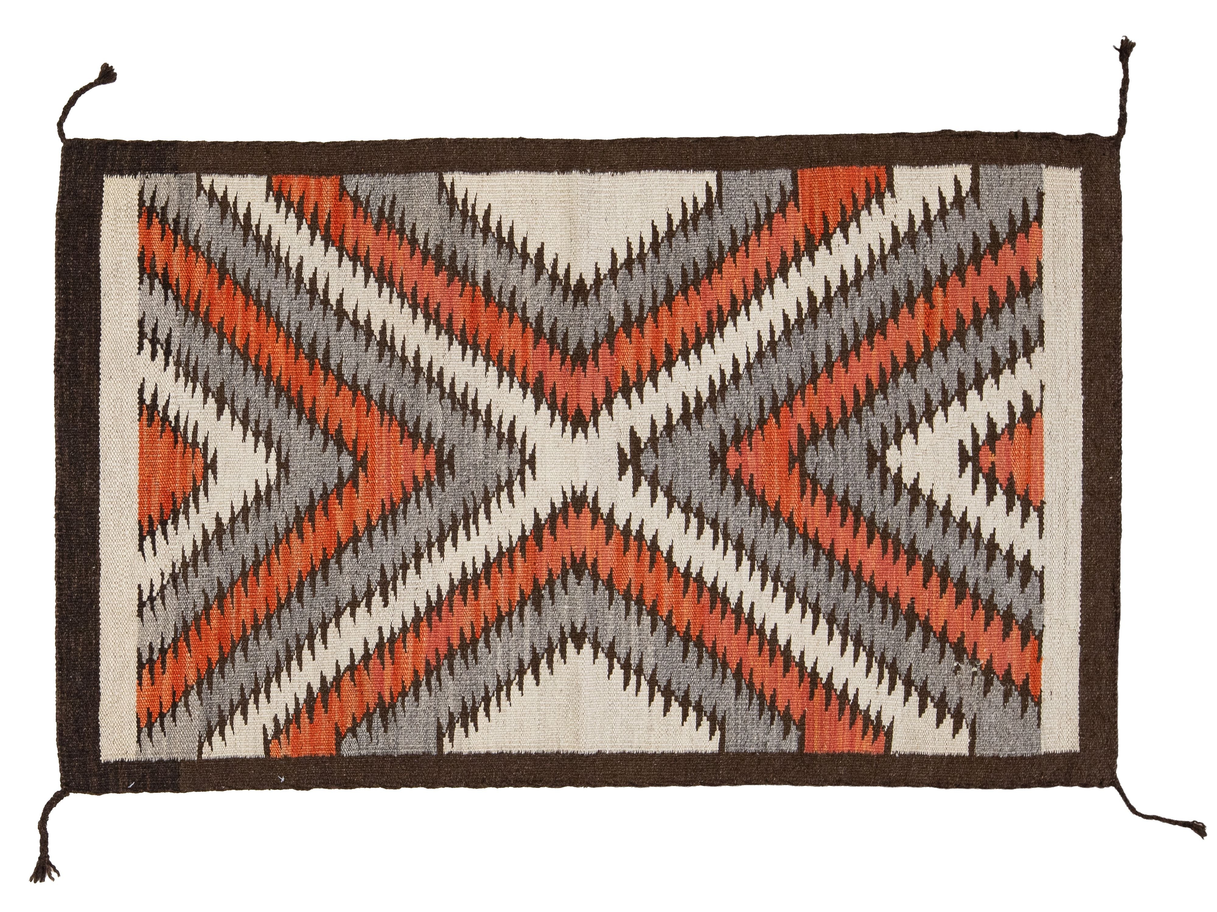 A rectangular woven textile rug or mat with a geometric Southwestern-style pattern in rust orange, gray, black, and off-white, finished with a dark brown border and hanging tassels at the corners.