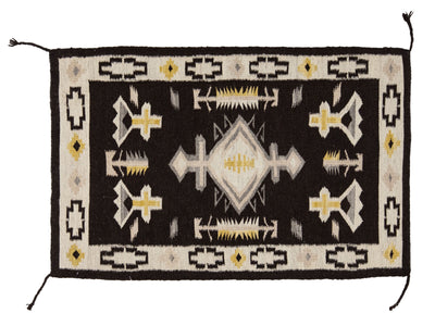 A rectangular Southwestern-style woven rug with a black center field, cream border, and geometric motifs including diamonds, crosses, and fish-like symbols in black, cream, gray, and mustard yellow.