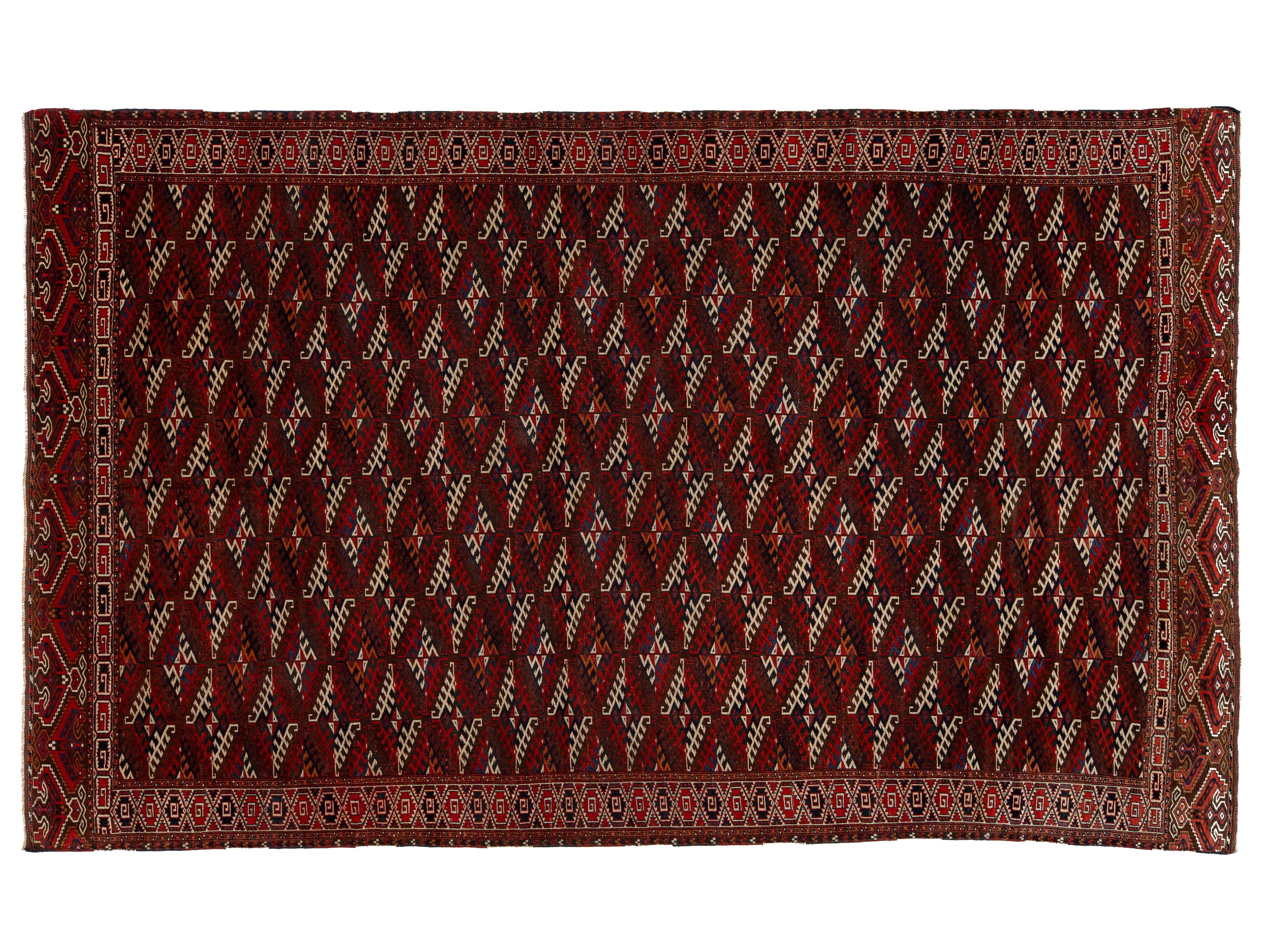 A rectangular traditional rug with a deep red field, featuring a repeating diamond pattern in cream, black, and red, and a detailed geometric border.