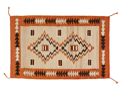 A rectangular woven rug with a Southwestern geometric pattern, featuring an orange border, a cream center, and black and orange zigzag diamond motifs.