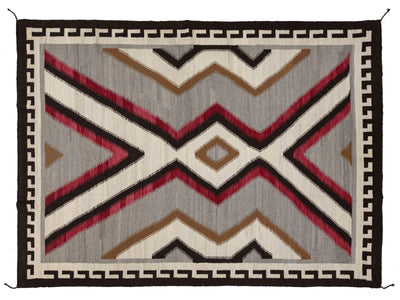 A rectangular woven rug with a Southwestern geometric pattern, featuring red, brown, black, and cream zigzags and diamonds on a gray background, finished with a black and cream Greek key border.