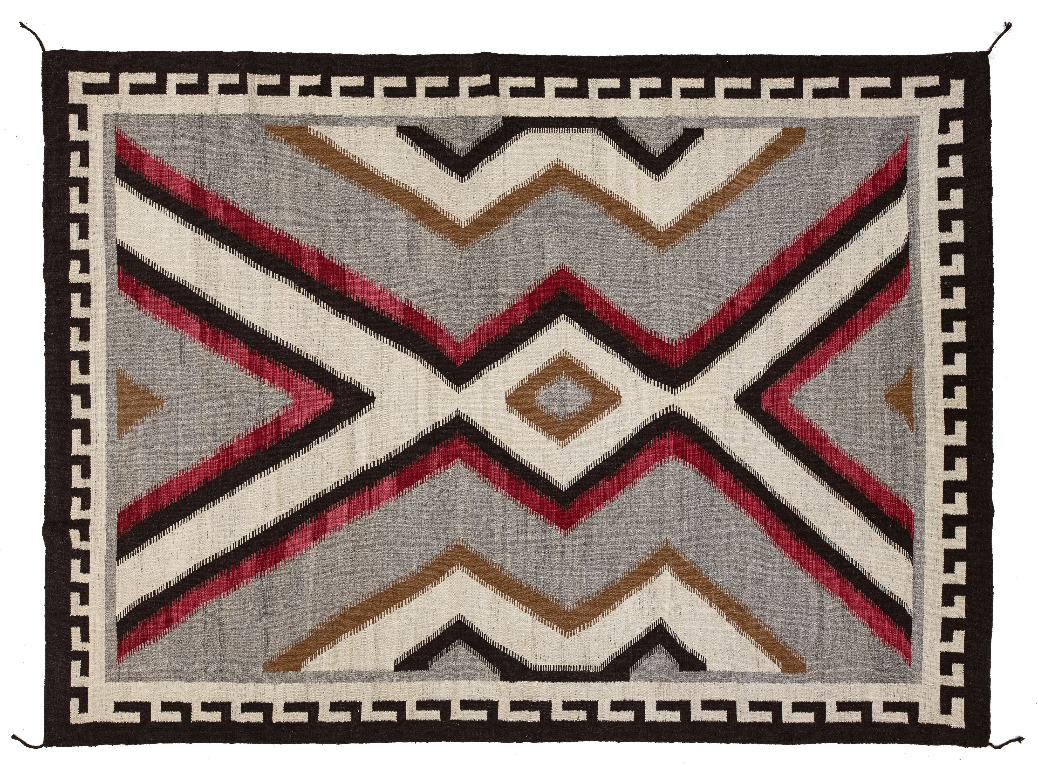 A rectangular woven rug with a Southwestern geometric pattern, featuring red, brown, black, and cream zigzags and diamonds on a gray background, finished with a black and cream Greek key border.
