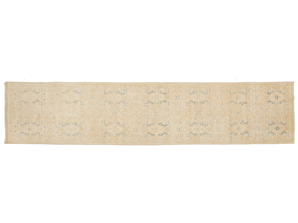 A runner rug in a light beige or cream tone with a faded, distressed pattern of muted blue and gray motifs.