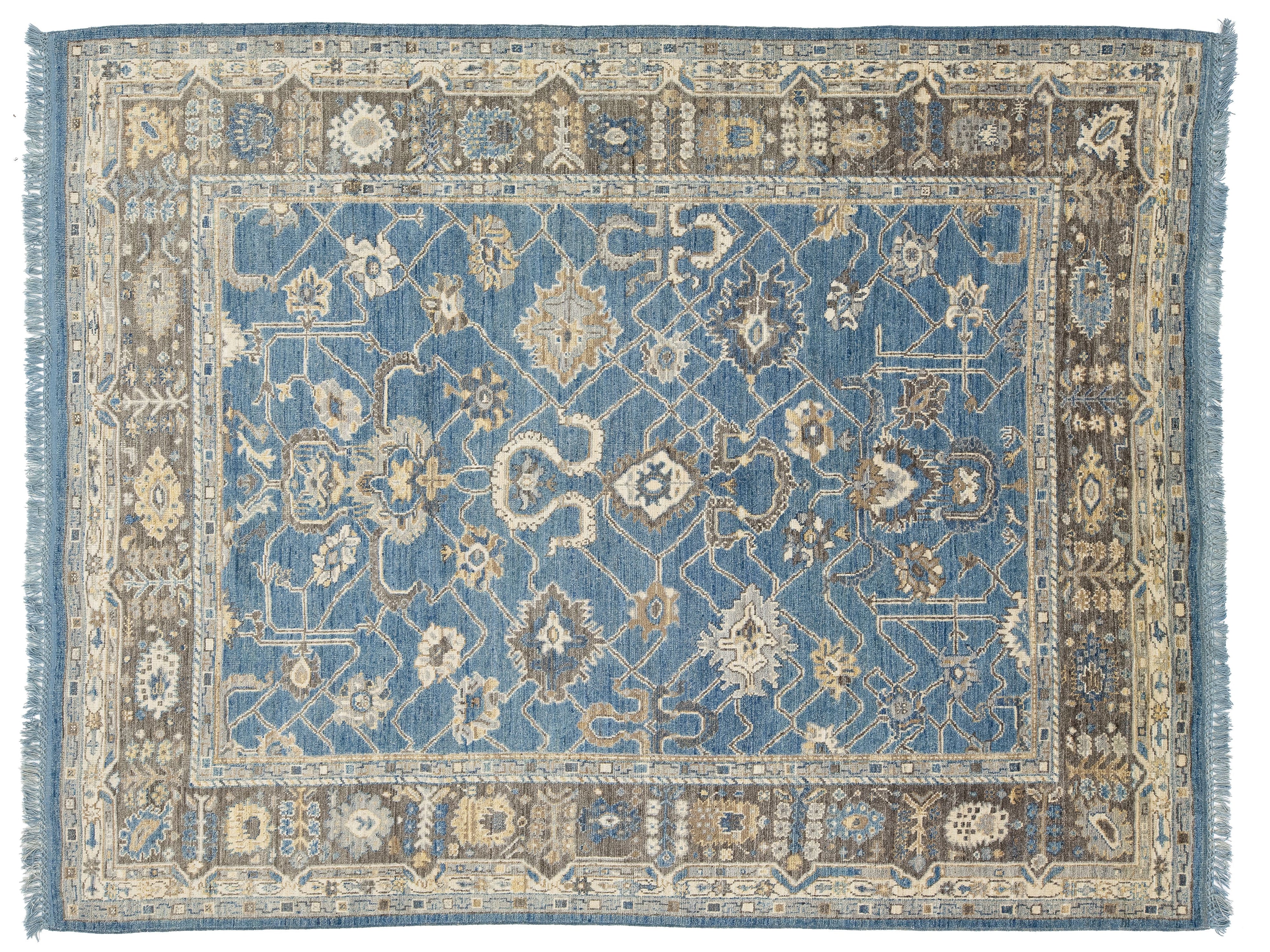 A rectangular traditional area rug with an intricate floral and geometric pattern, featuring a dominant medium blue center field, a beige and brown border, and blue fringe on the sides.