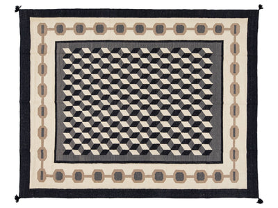 A rectangular woven rug with a bold geometric pattern, featuring a central black and cream 3D cube illusion pattern, surrounded by a cream field with taupe chain motifs and a solid black outer border.