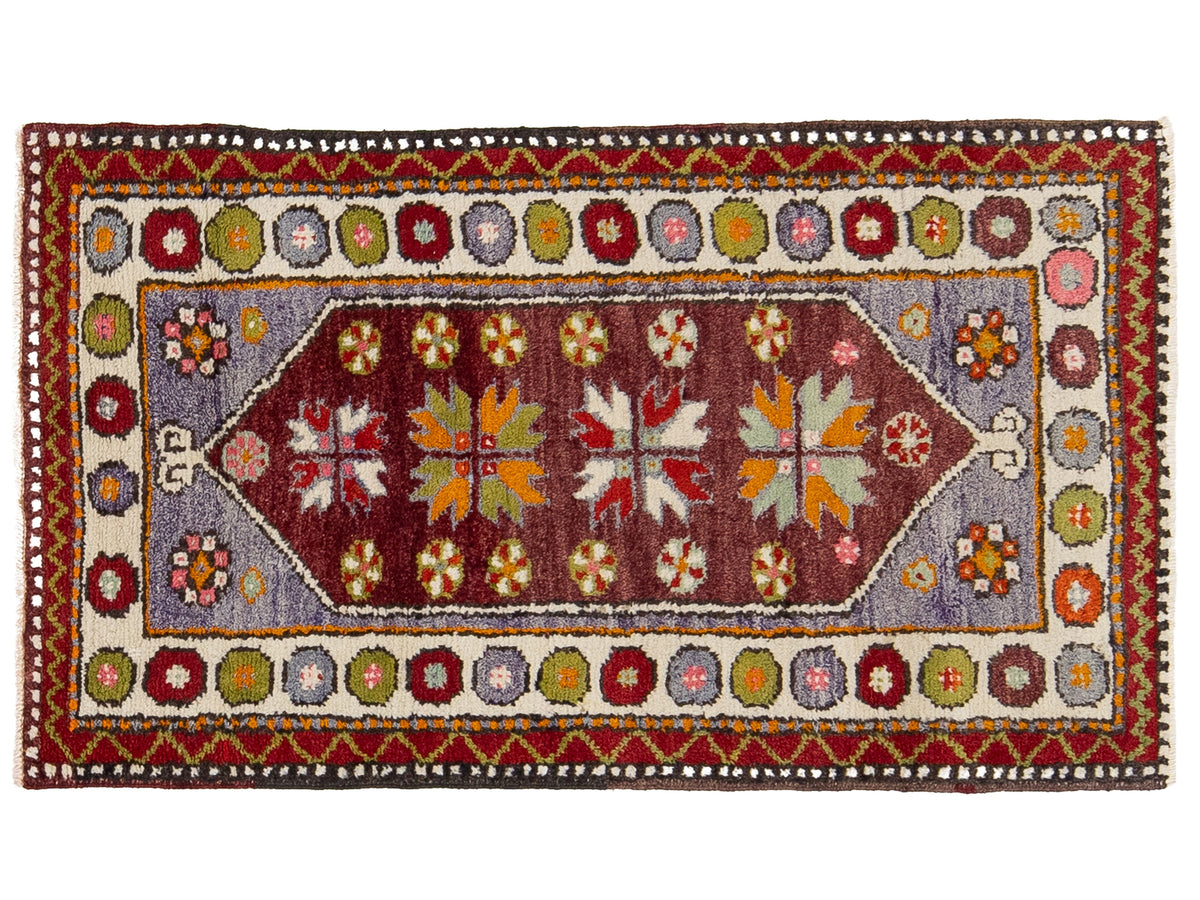 A traditional hand-knotted wool rug with a geometric pattern, featuring a central red field, a light blue-gray center panel, and a cream-colored border with red, green, and blue accents.