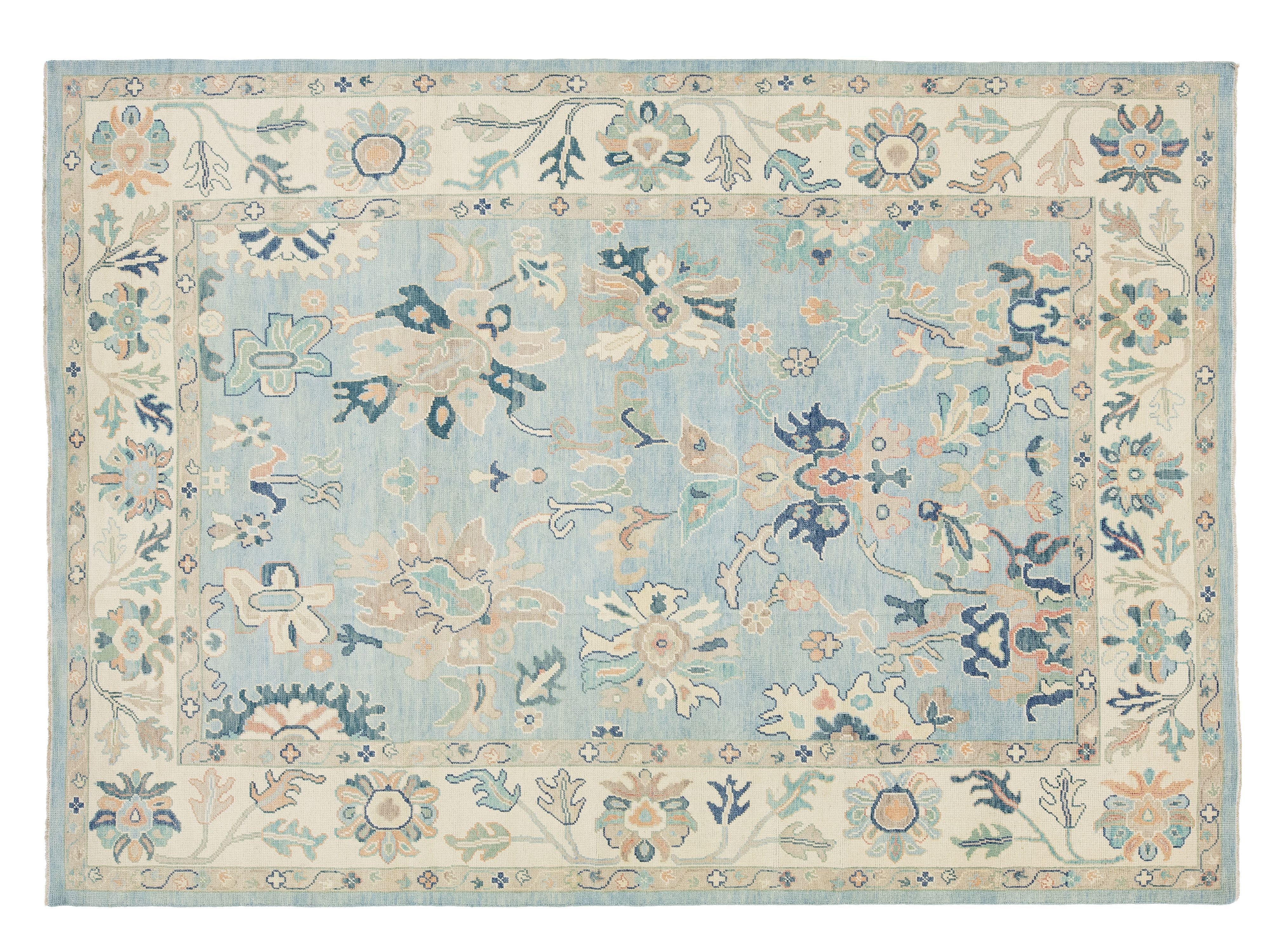 A rectangular area rug with an intricate floral and botanical pattern, featuring a light blue center field and a cream border with blue, green, and peach details.