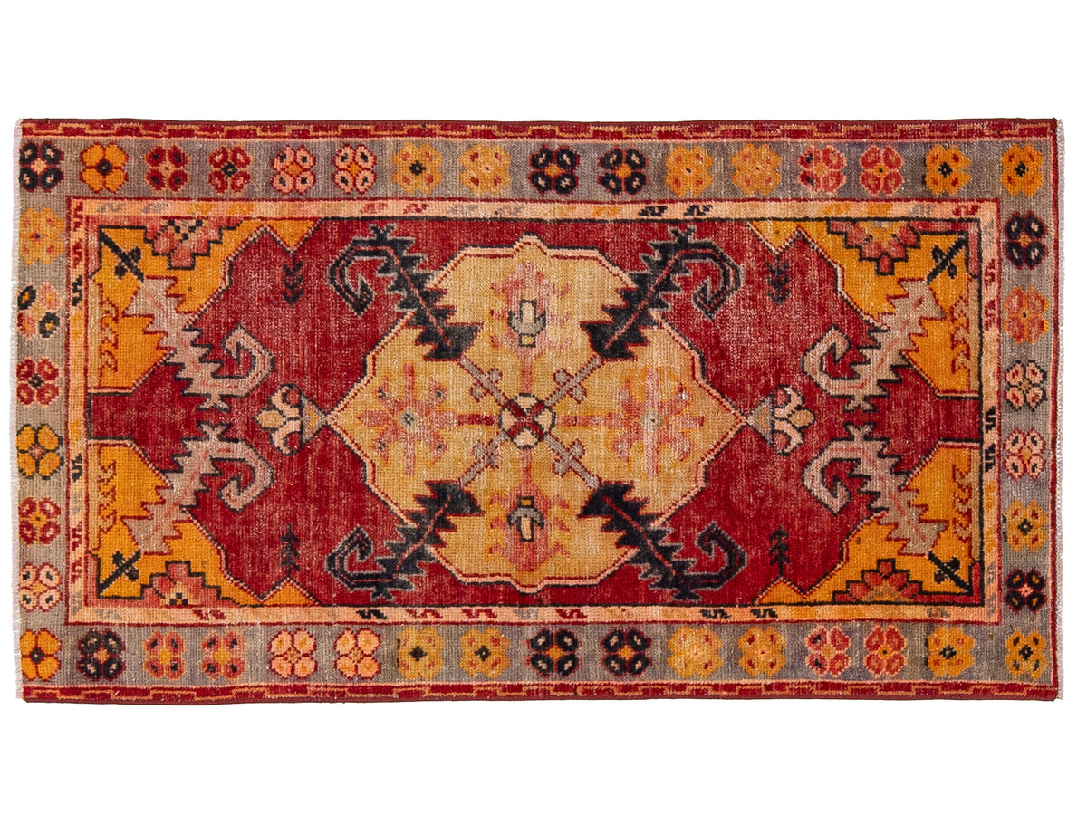 A rectangular vintage area rug with a distressed geometric pattern in red, orange, beige, and gray, featuring a central medallion and decorative borders.