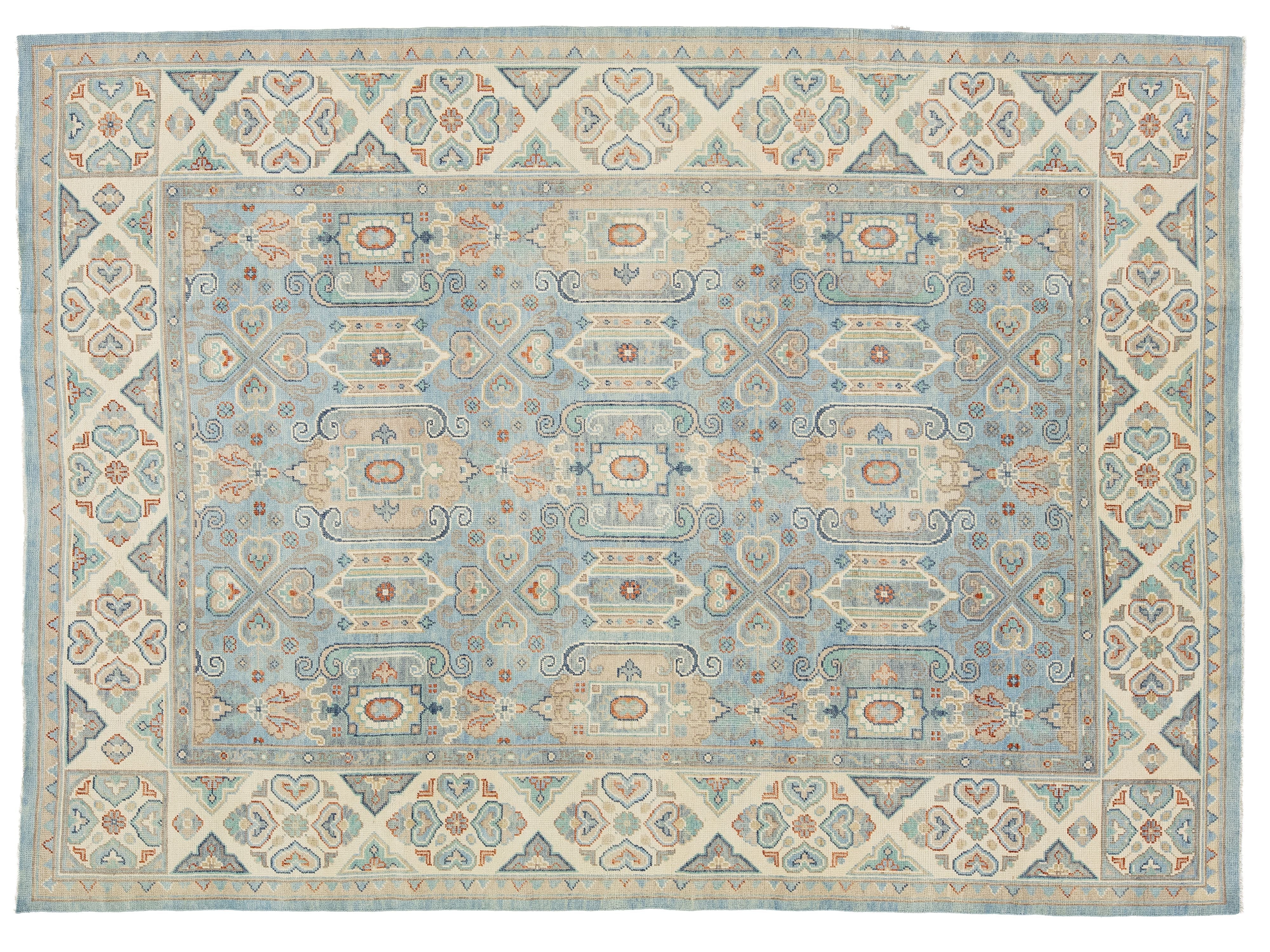 A rectangular area rug with a traditional ornamental pattern, featuring a light blue center field, beige borders, and accents of rust red, navy, and gray.