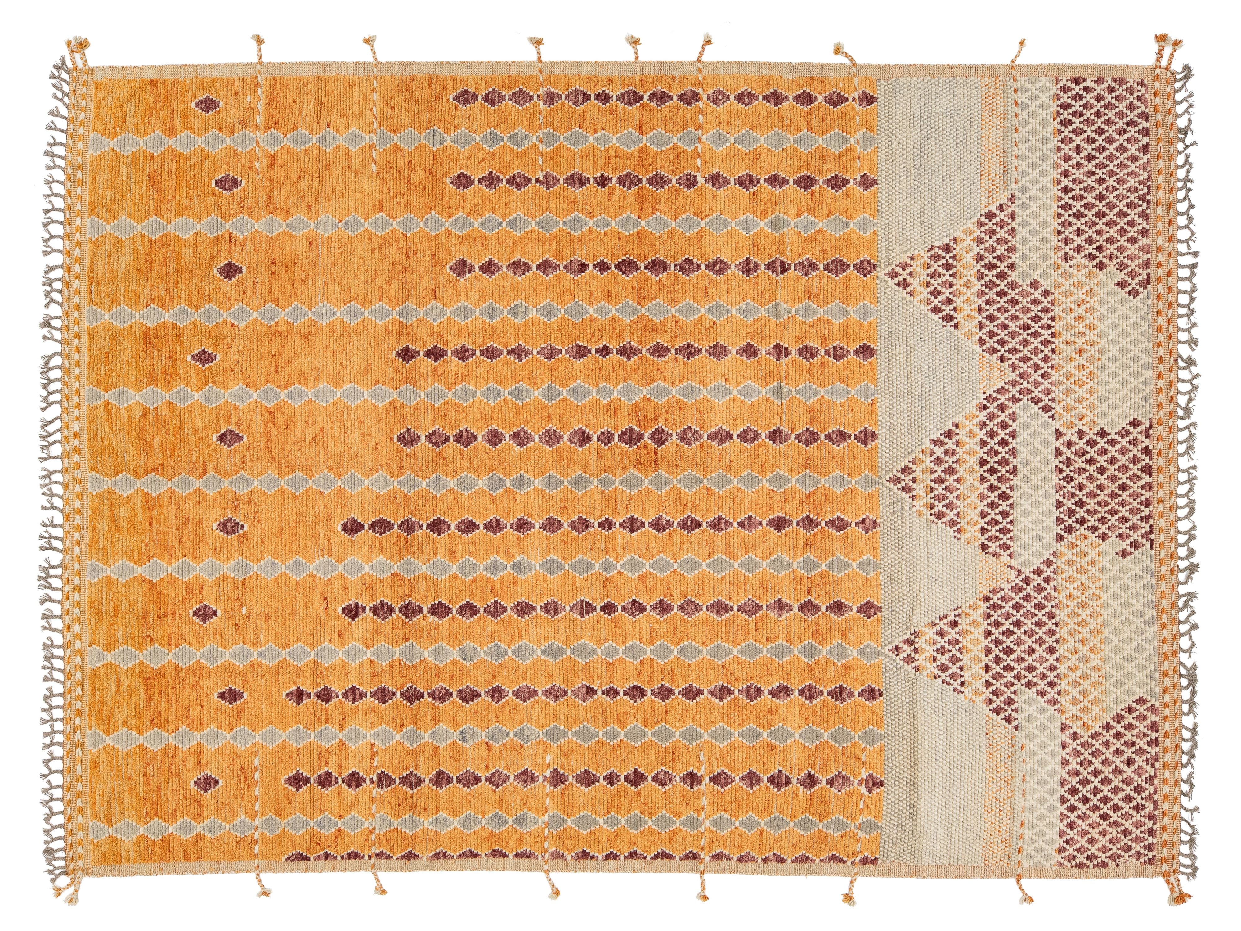 A rectangular woven rug with a warm ochre-orange field, horizontal bands of small dark red and beige diamond motifs, and a patterned border in beige, rust, and cream with fringe tassels on the sides.