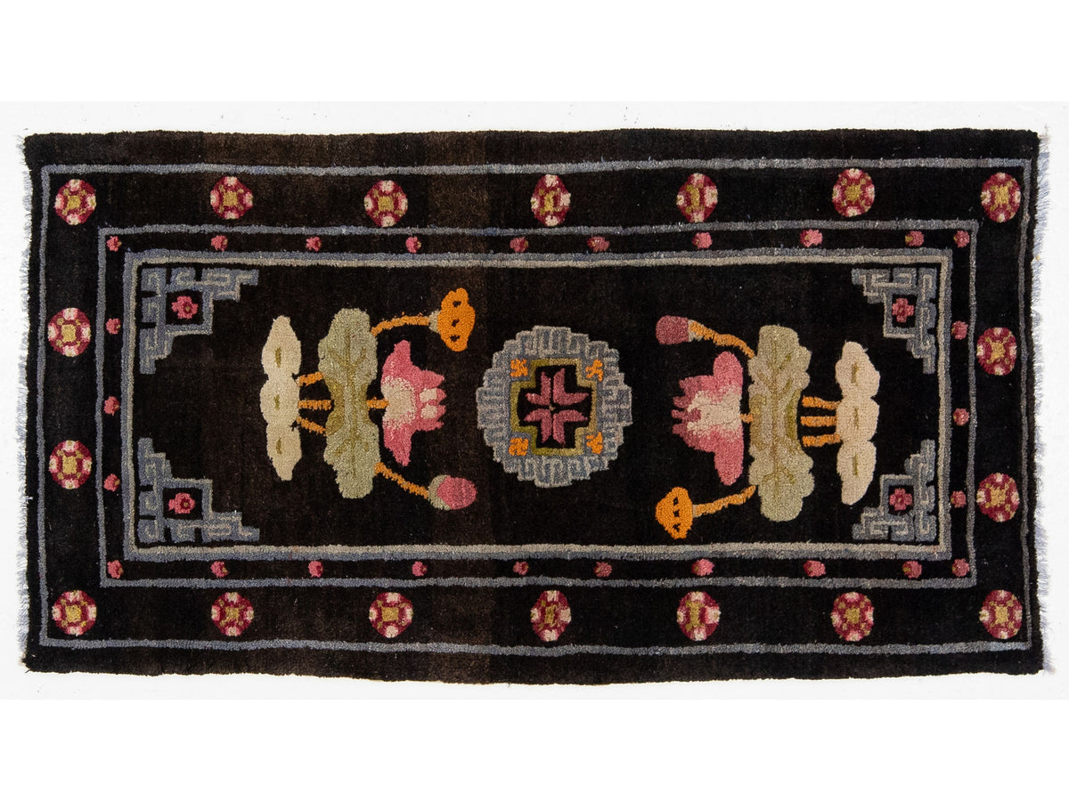 A rectangular hand knotted rug with a black field, featuring a central medallion and lotus motifs in pink, beige, orange, and gray, surrounded by a decorative border with pink and orange floral accents.