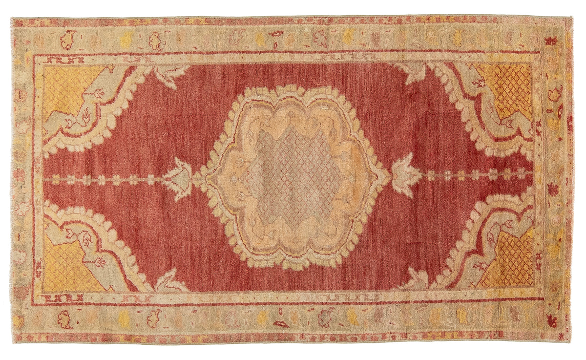 A rectangular vintage area rug with a central medallion design in muted red, beige, and ochre yellow tones, shown flat against a white background.
