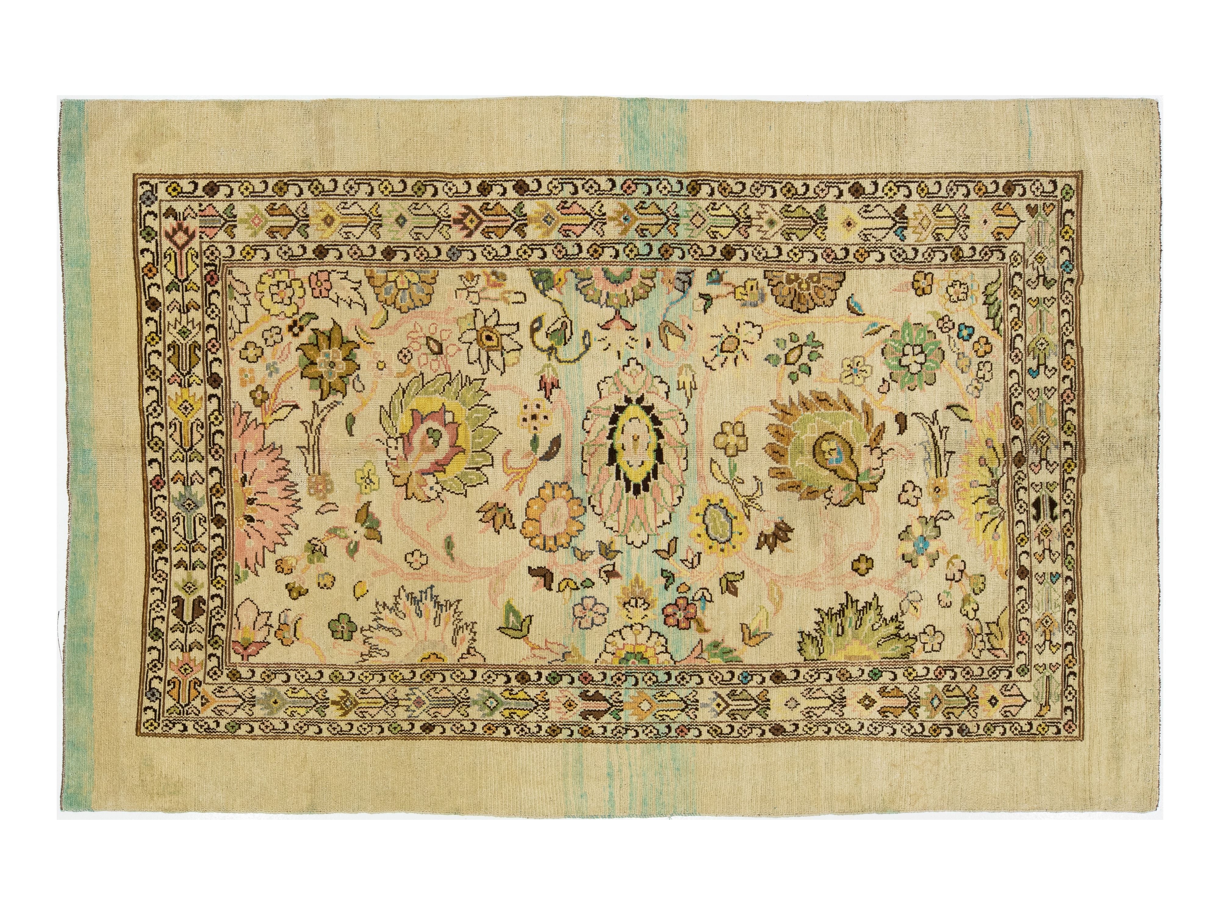 Hand-knotted wool rug with a cream-colored field, featuring a central floral medallion and a detailed border in muted tones of green, pink, yellow, and brown.