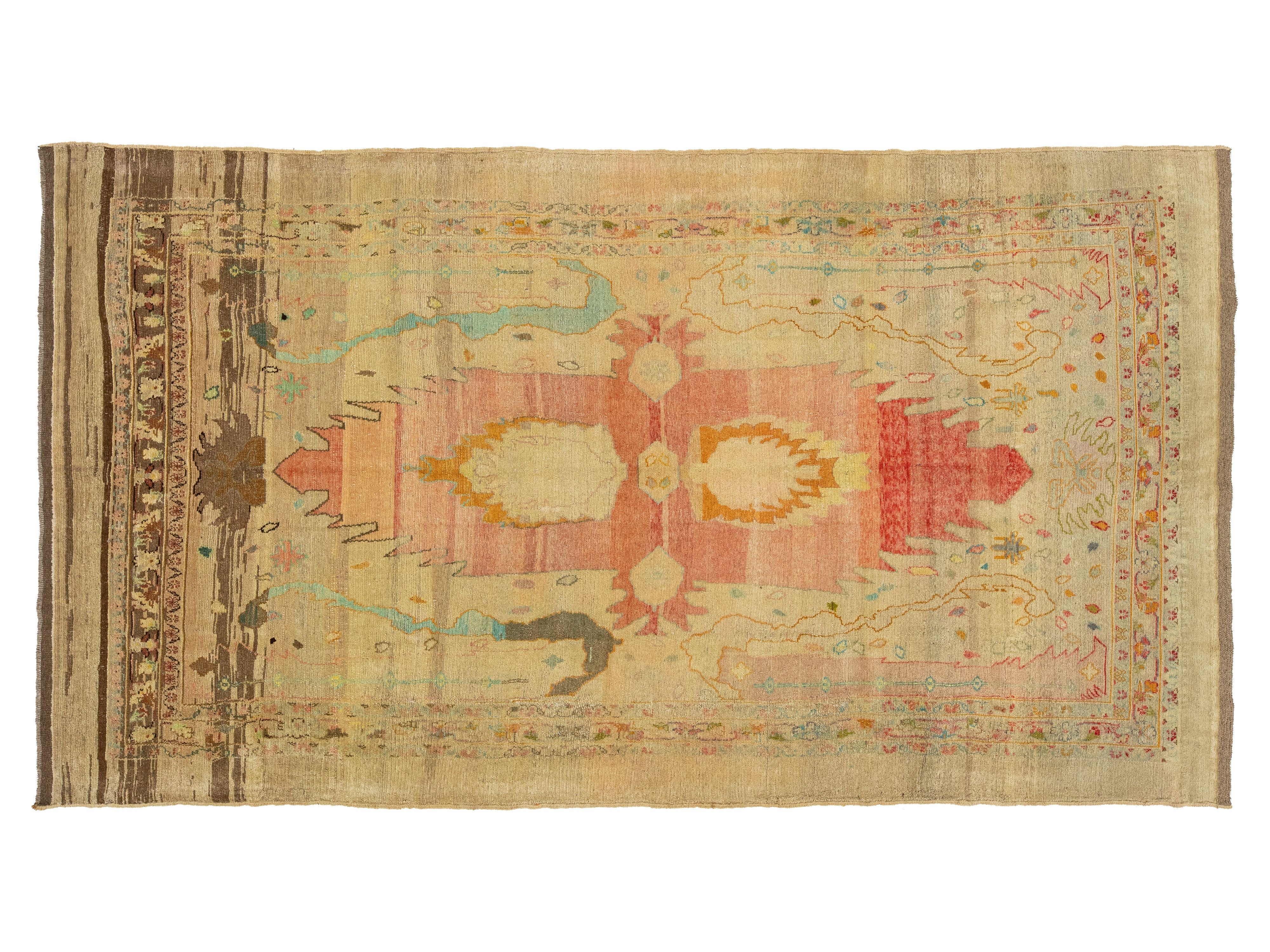A flat-laid vintage-style area rug with a distressed, hand-knotted texture. The rug features a central medallion motif in muted shades of pink, ochre yellow, and light blue on a warm beige background, framed by a brown patterned border on the left side.