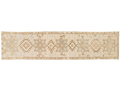 A runner rug with a cream and beige base and repeating geometric medallion motifs in tan, muted blue, and brown.
