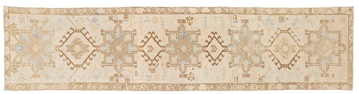 A runner rug with a cream and beige base and repeating geometric medallion motifs in tan, muted blue, and brown.