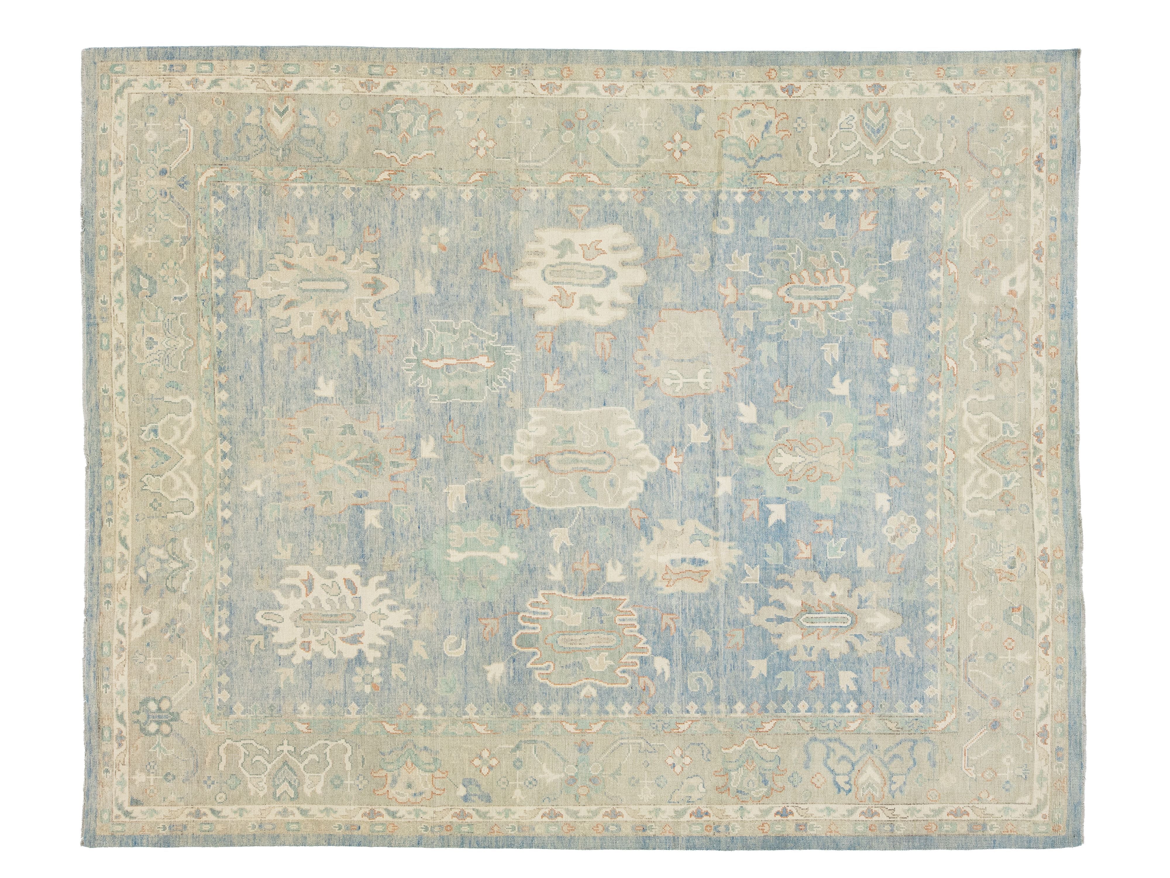 A rectangular area rug with a traditional design, featuring a dusty blue center field, beige borders, and muted floral medallions in cream, sage green, and light orange.