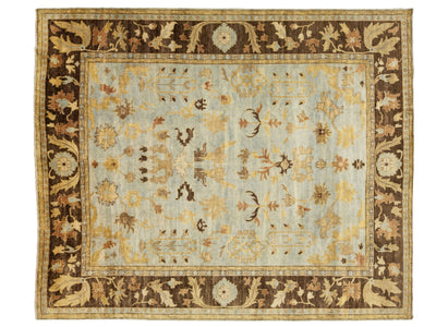 A rectangular traditional area rug with a light blue-gray field, a wide dark brown border, and intricate floral motifs in beige, mustard, and brown.