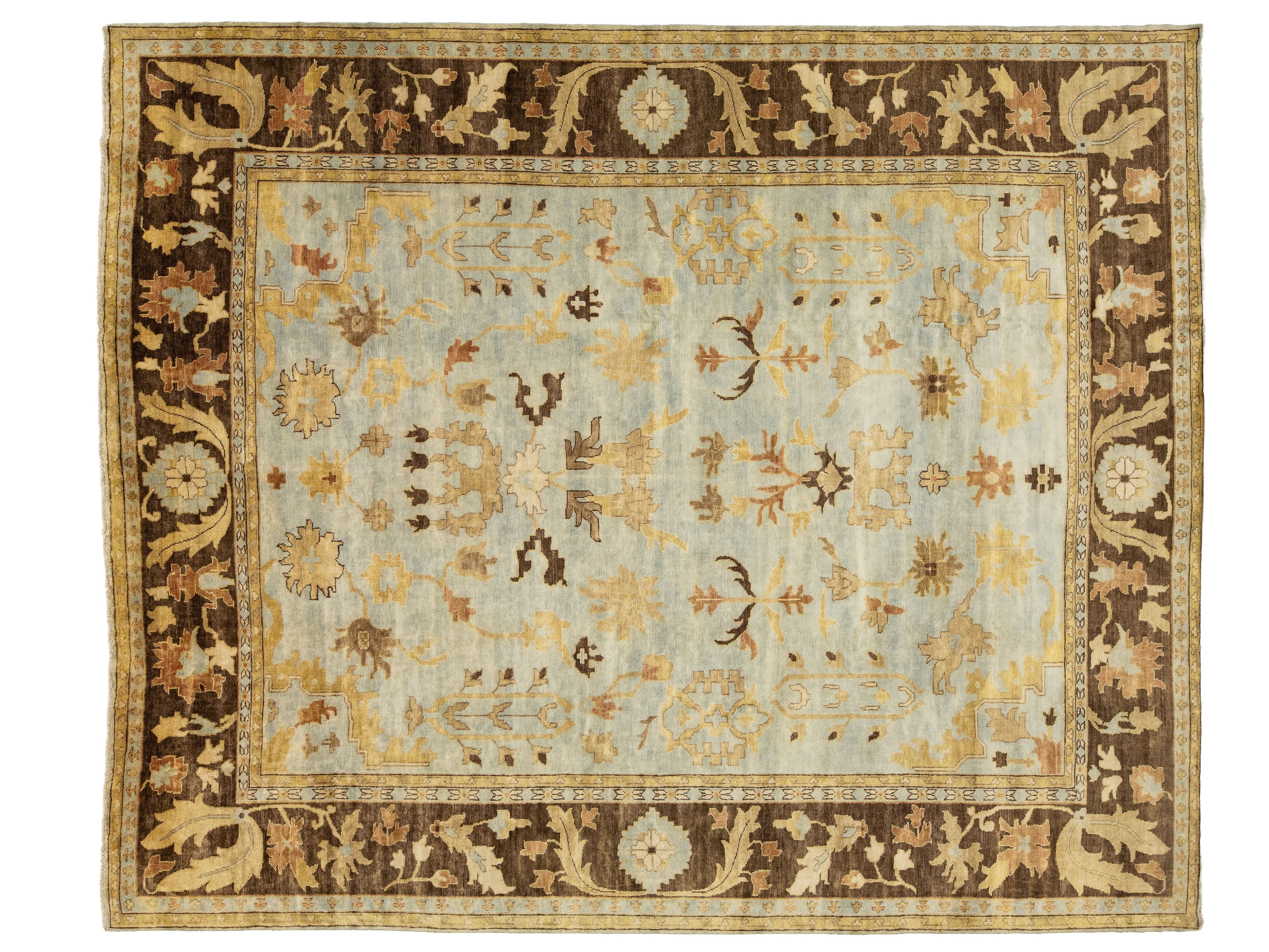A rectangular traditional area rug with a light blue-gray field, a wide dark brown border, and intricate floral motifs in beige, mustard, and brown.