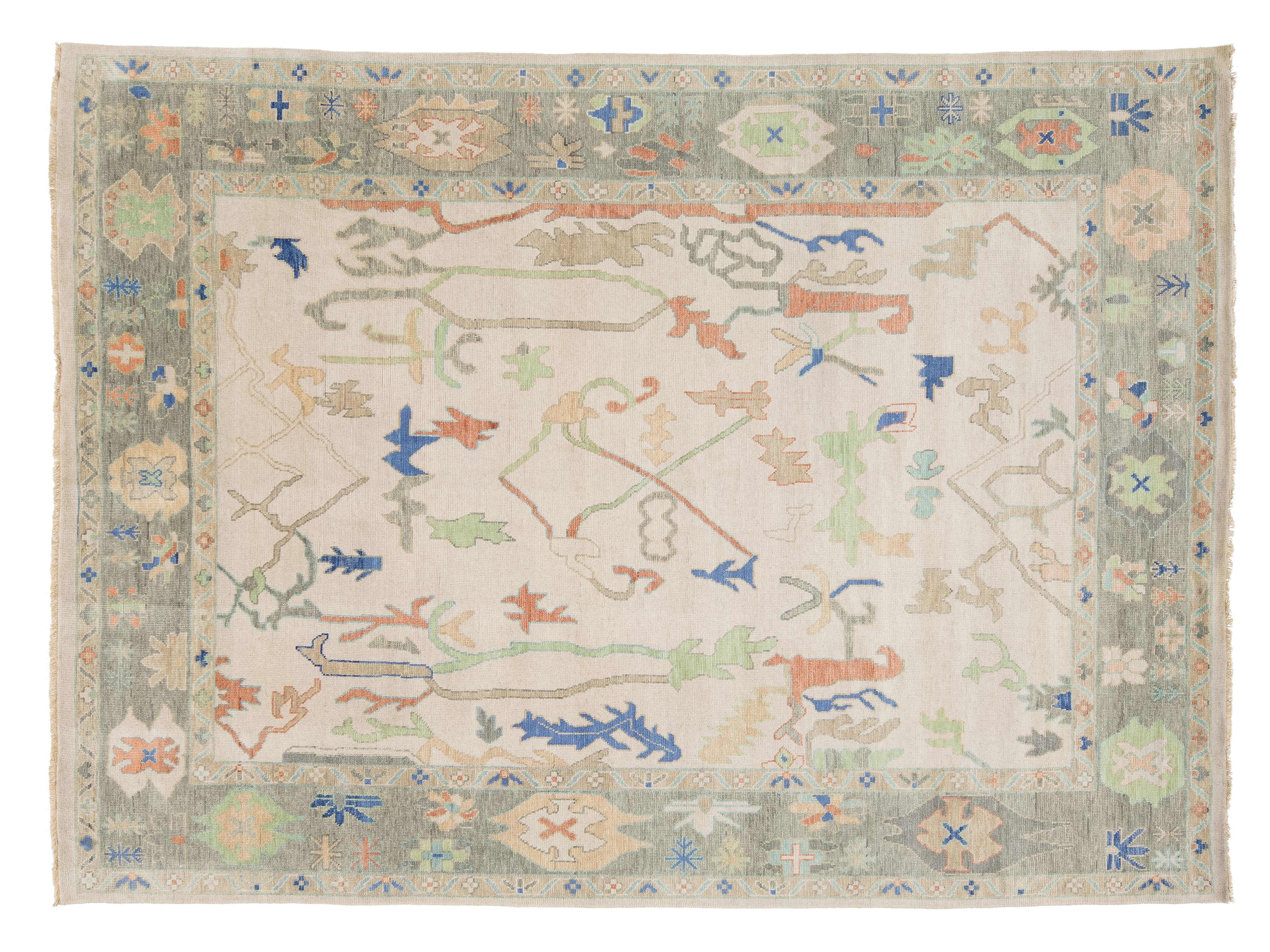 A rectangular hand-knotted rug with a cream center field, a muted sage green border, and scattered abstract motifs in blue, rust orange, and light green.