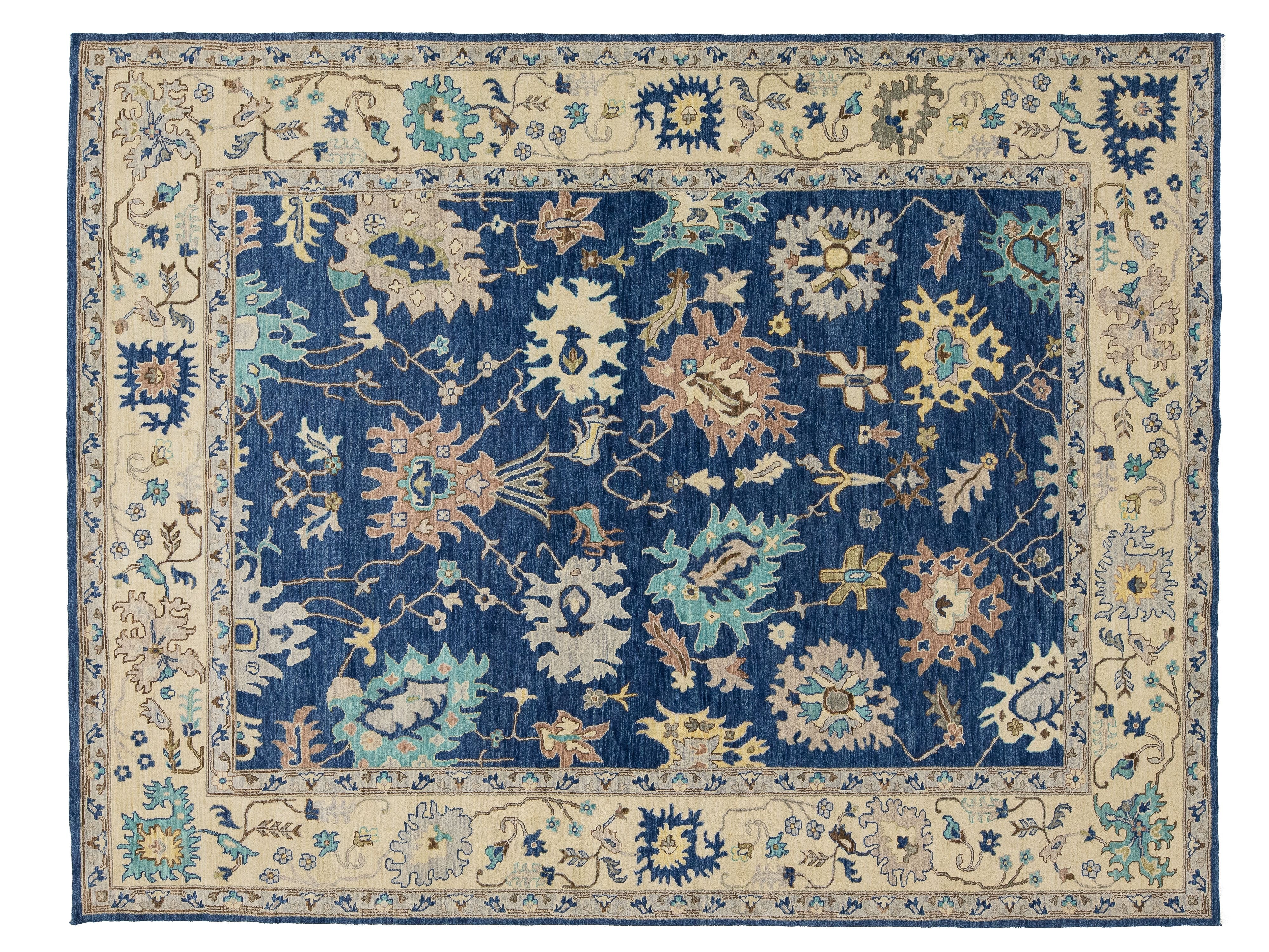A rectangular traditional area rug with a navy blue central field, surrounded by a beige border and detailed floral motifs in blue, beige, and light teal.