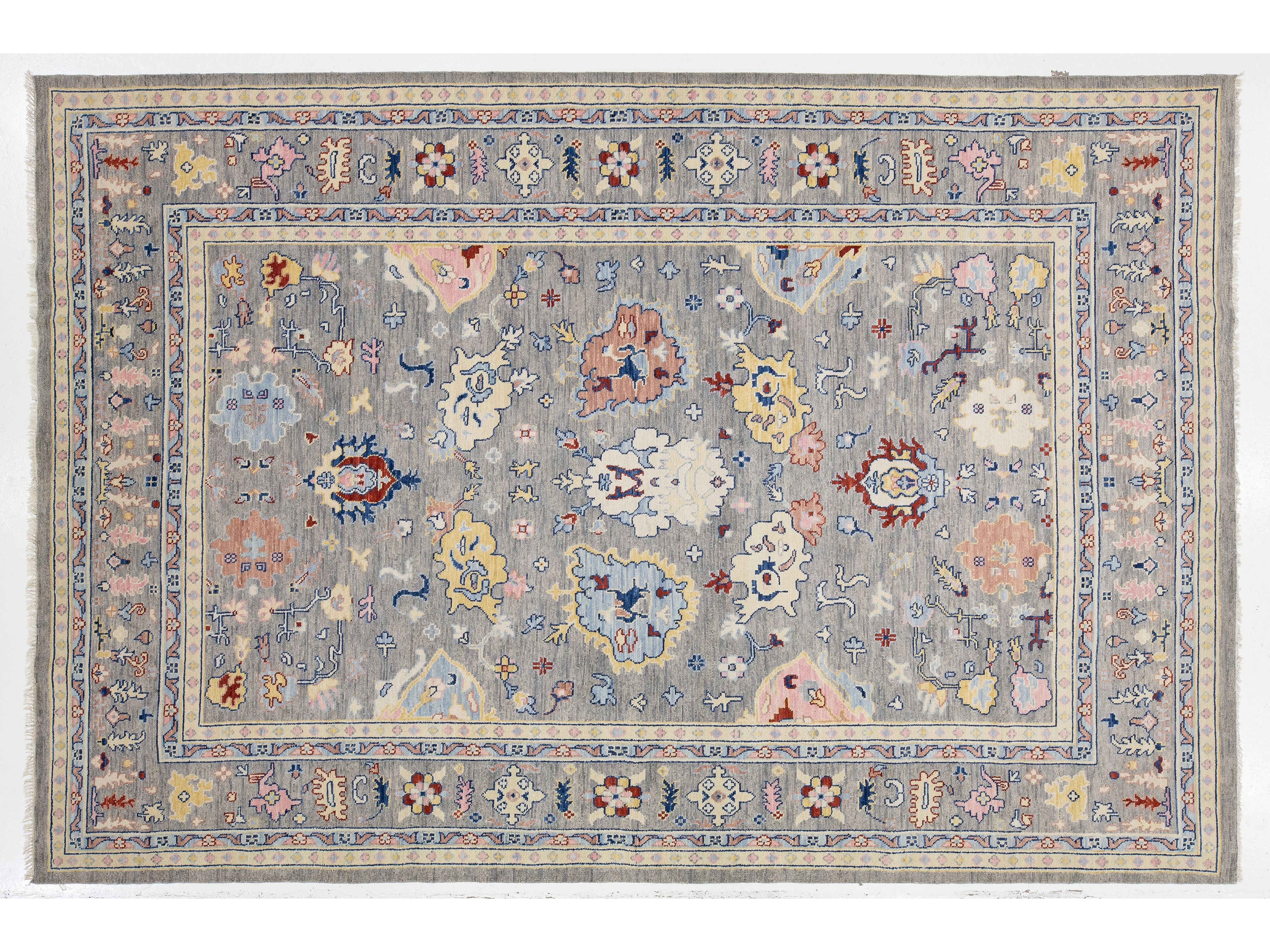 A rectangular area rug with an intricate traditional floral pattern, featuring a light gray-blue field and a detailed border in muted shades of pink, blue, beige, and red.