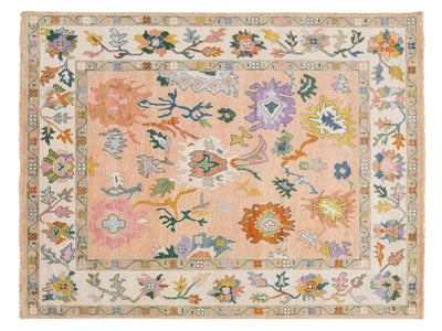 A rectangular area rug with a traditional hand-knotted design, featuring a dusty pink center field surrounded by a cream-colored border with multicolored floral motifs.