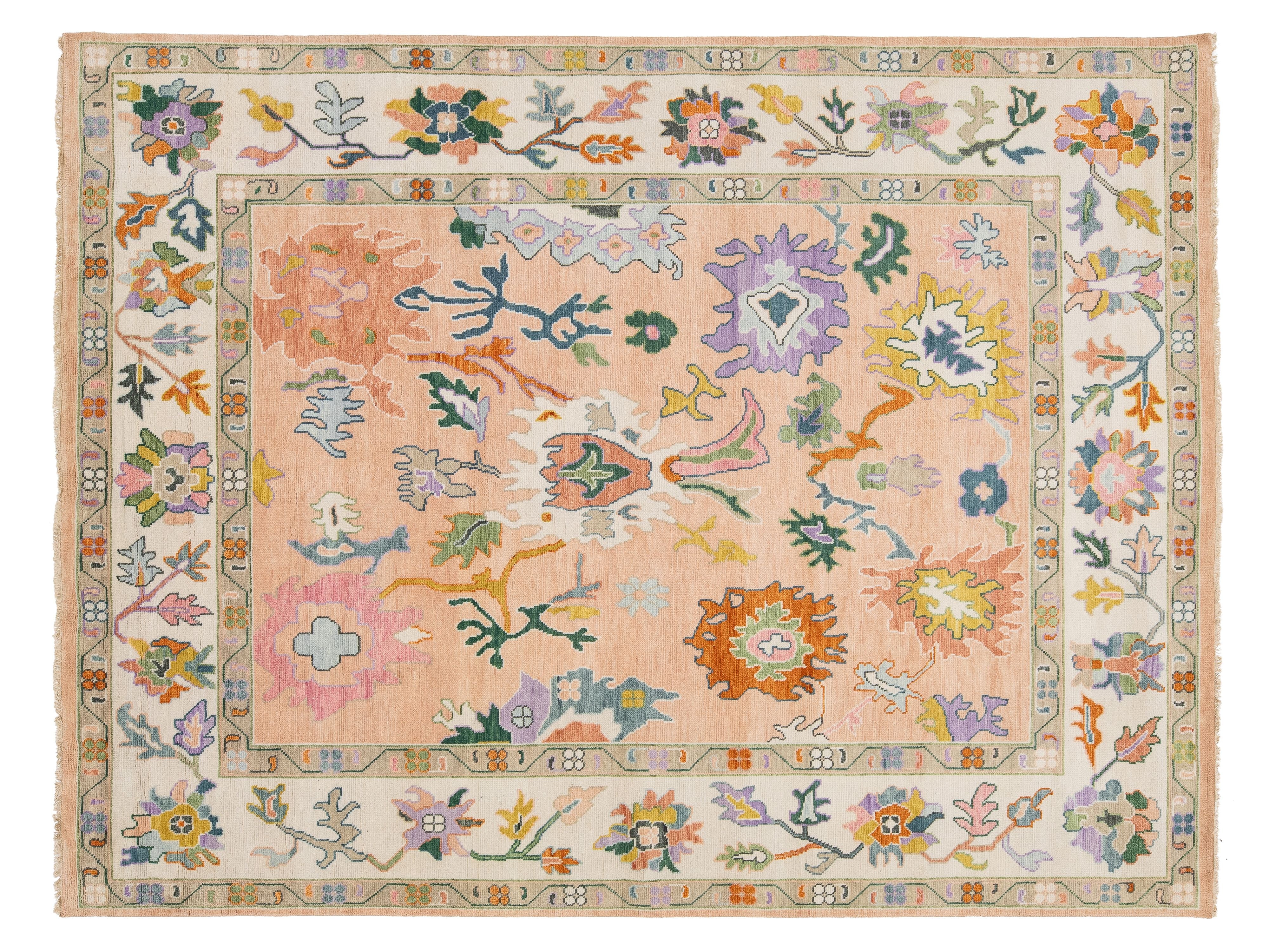 A rectangular area rug with a traditional hand-knotted design, featuring a dusty pink center field surrounded by a cream-colored border with multicolored floral motifs.