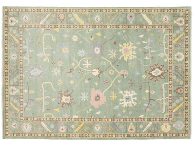 A rectangular area rug with a muted sage green field, featuring a detailed traditional pattern in soft pastel tones of pink, beige, yellow, and brown, surrounded by a decorative border.