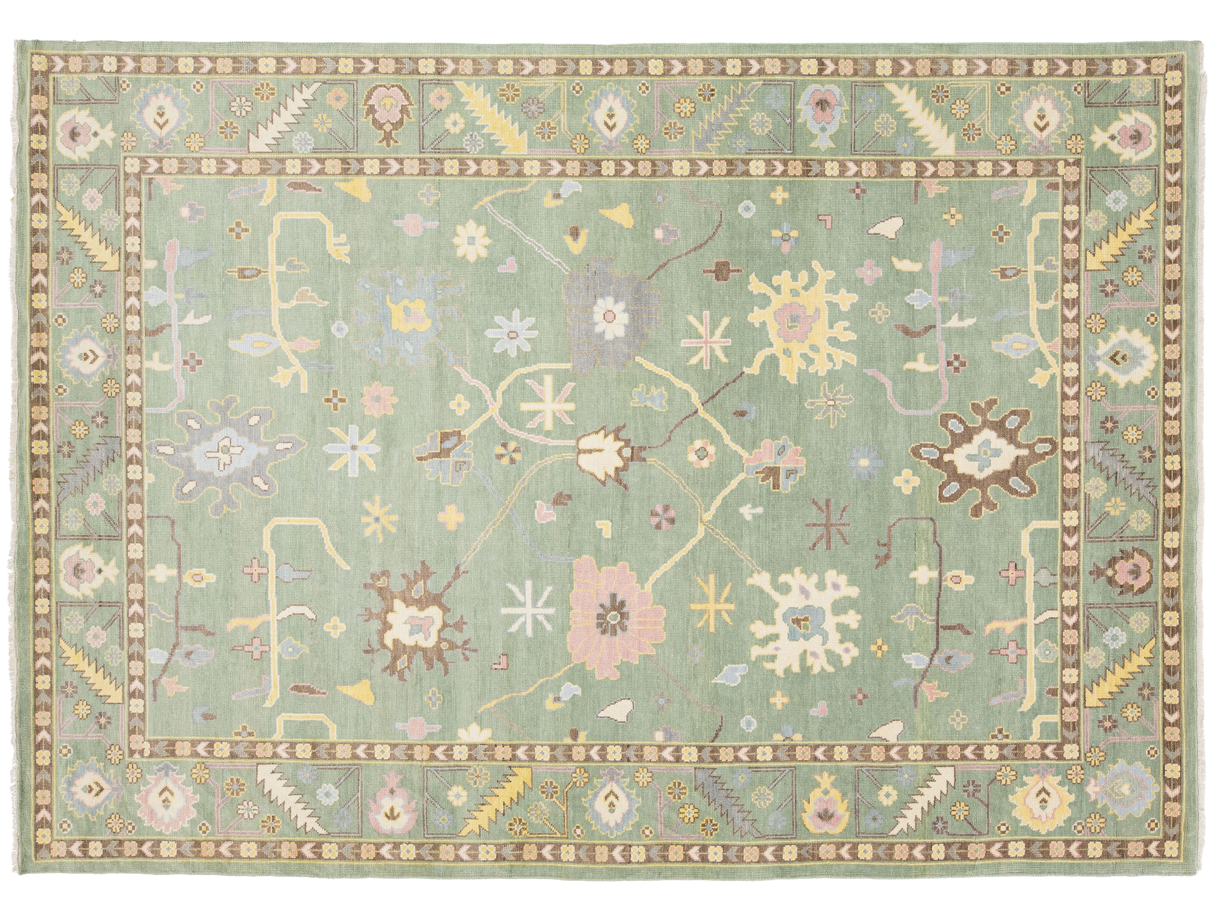 A rectangular area rug with a muted sage green field, featuring a detailed traditional pattern in soft pastel tones of pink, beige, yellow, and brown, surrounded by a decorative border.