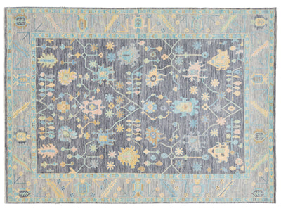 A rectangular area rug with a distressed traditional pattern, featuring a central field of charcoal gray with light blue, beige, and yellow floral motifs, surrounded by a light blue border.