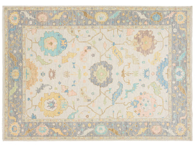 A rectangular area rug with an intricate traditional floral pattern in soft pastel colors including blue, yellow, pink, and green on a cream background, surrounded by a dusty blue border.