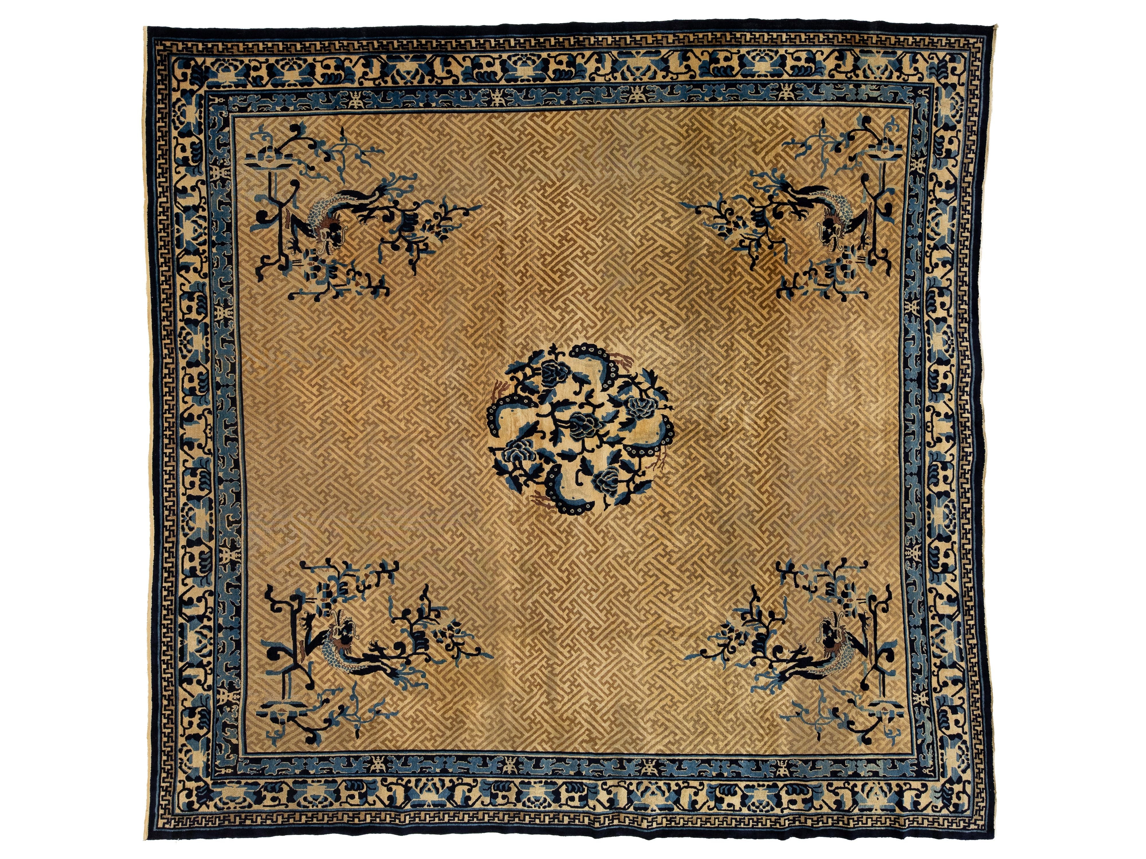 A square antique oriental rug with a tan and gold background, featuring a central floral medallion, four corner floral motifs, and a detailed blue and black border.